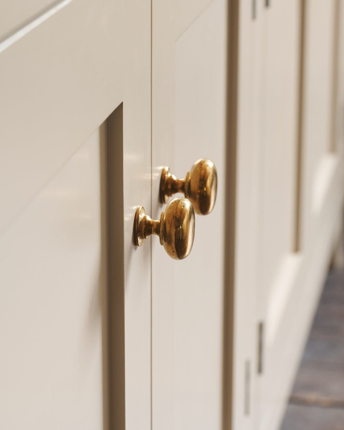 A close-up profile view of a pair of classic knobs in a warm burnished brass finish, fitted to a mushroom painted Shaker-style kitchen cupboard. TEST
