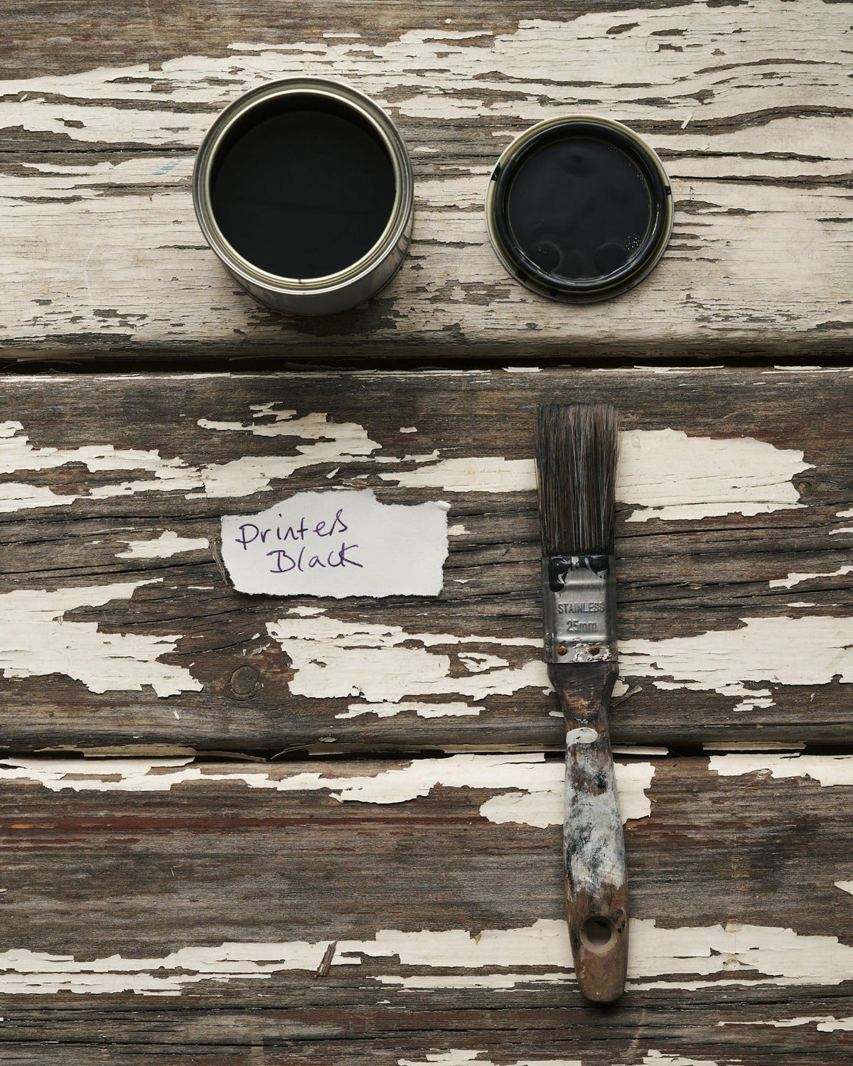 An overhead view of an open paint pot and a worn brush on a rustic tabletop. The black colour inside is labelled 'Printer's Black'. TEST
