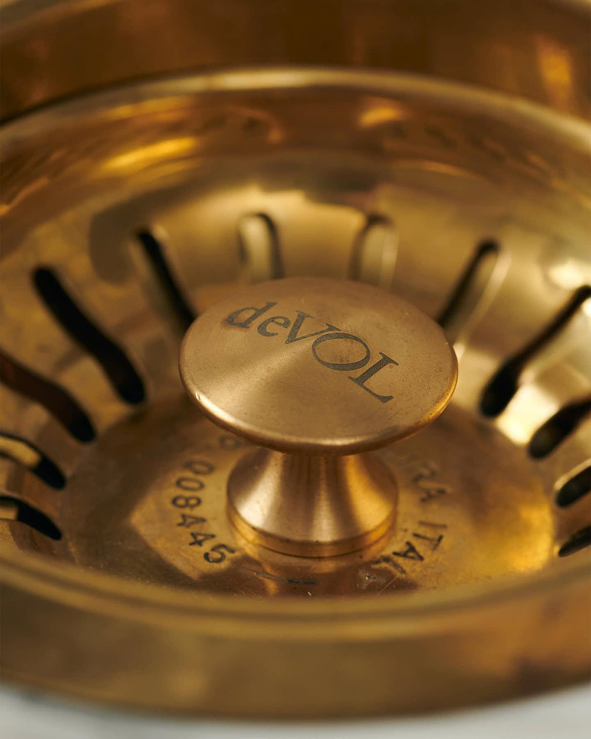 A close-up view of the central finial of a warm burnished brass basket waste strainer, there is a small 'deVOL' logo in the middle of the finial. TEST