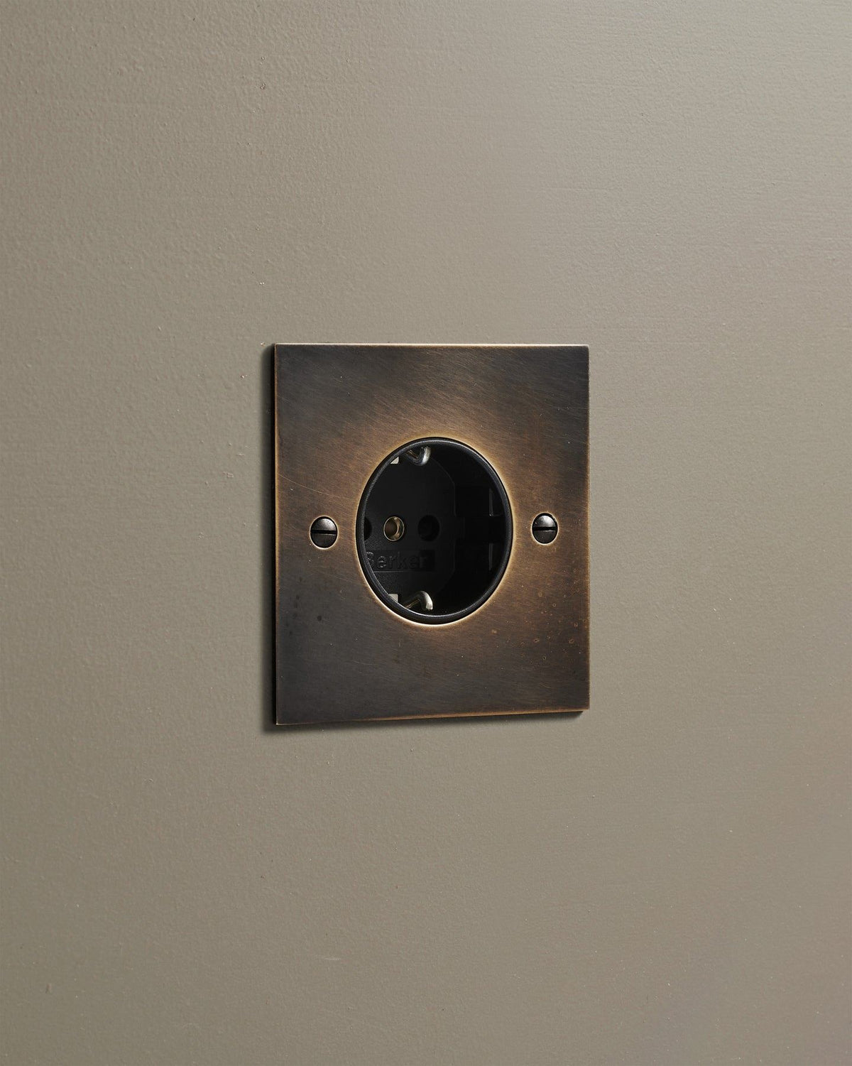 An oblique view of a classic Schuko-style single plug socket in a dark oxidised brass finish. TEST