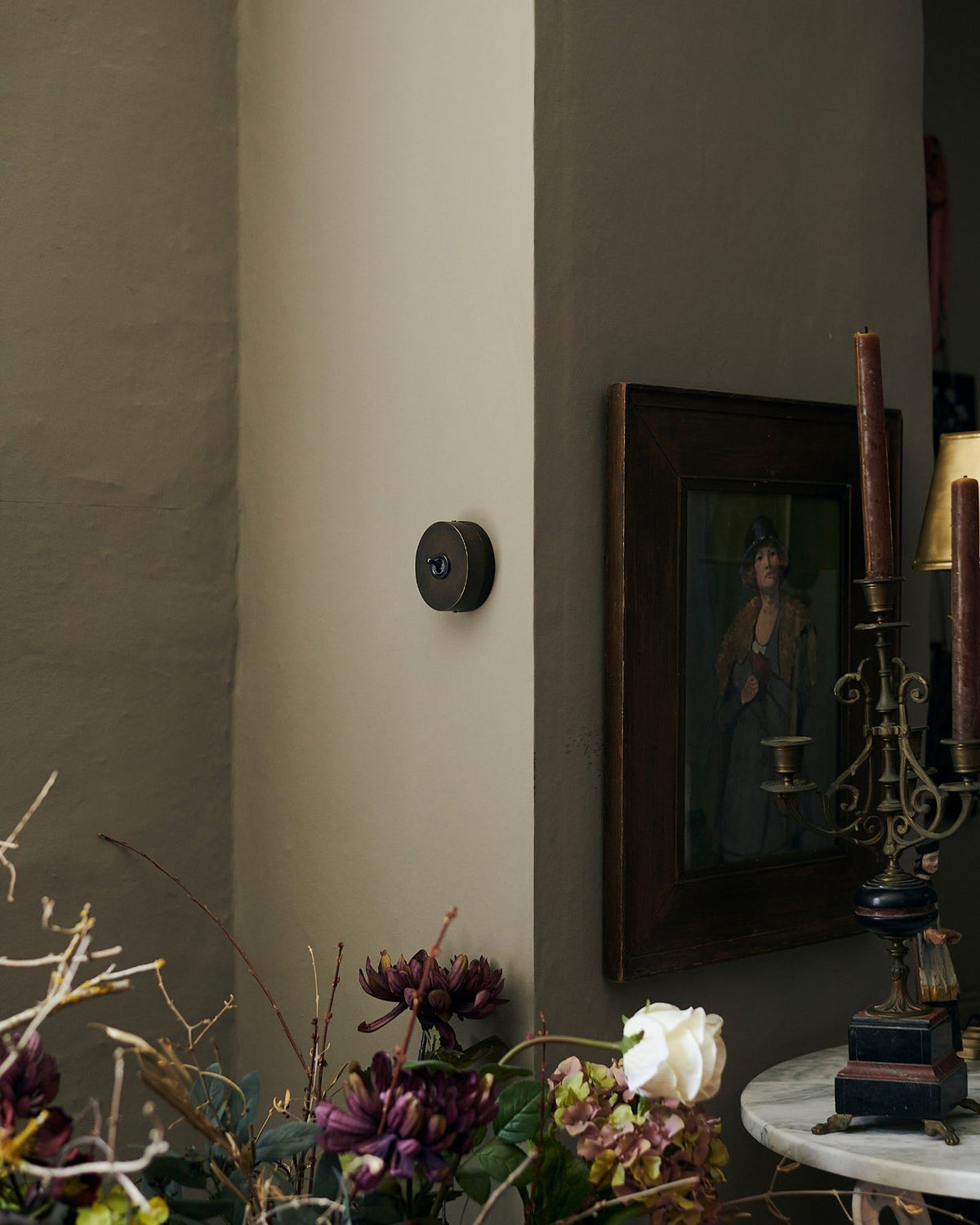 A small 1 gang oval-shaped toggle switch in a dark oxidised brass finish on a grey wall, a moody impressionist oil painted hangs on the wall around a corner with dried flowers in the foreground. | 1 Gang shown TEST