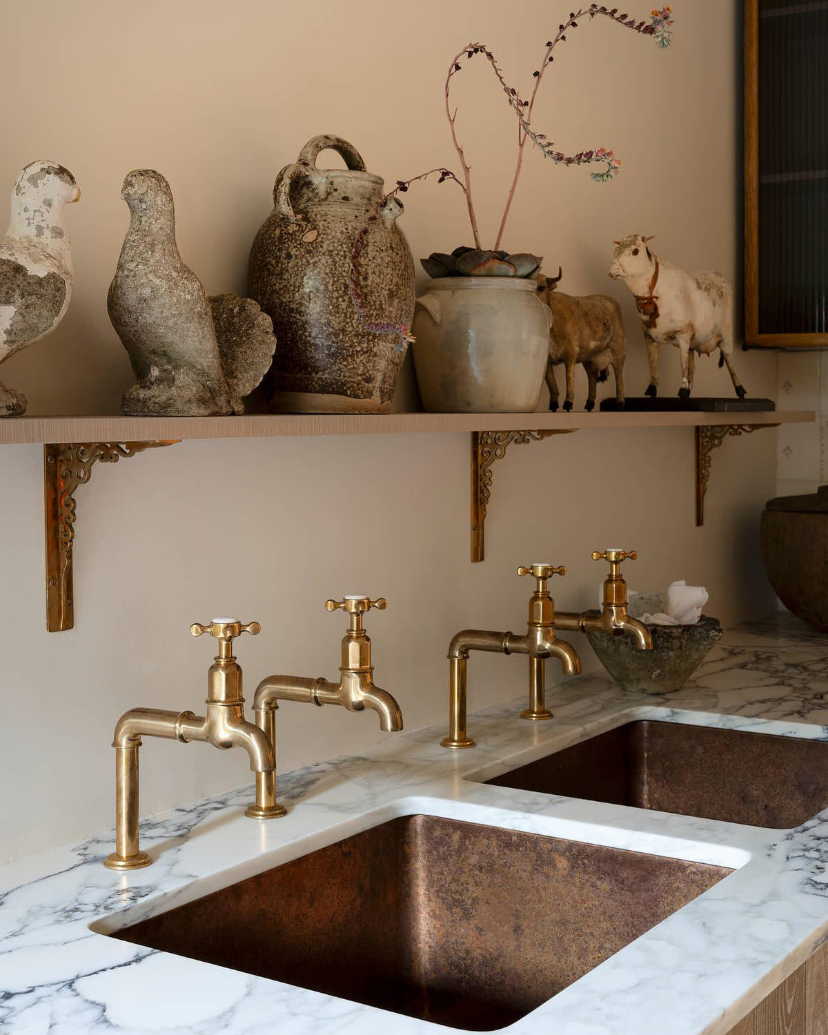 A close-up view of a pair of traditional-style aged brass bibcock taps with crosshead handles. An 'H' can be seen in the centre of the handle with the words 'deVOL' and 'Made in England' around the perimeter. TEST