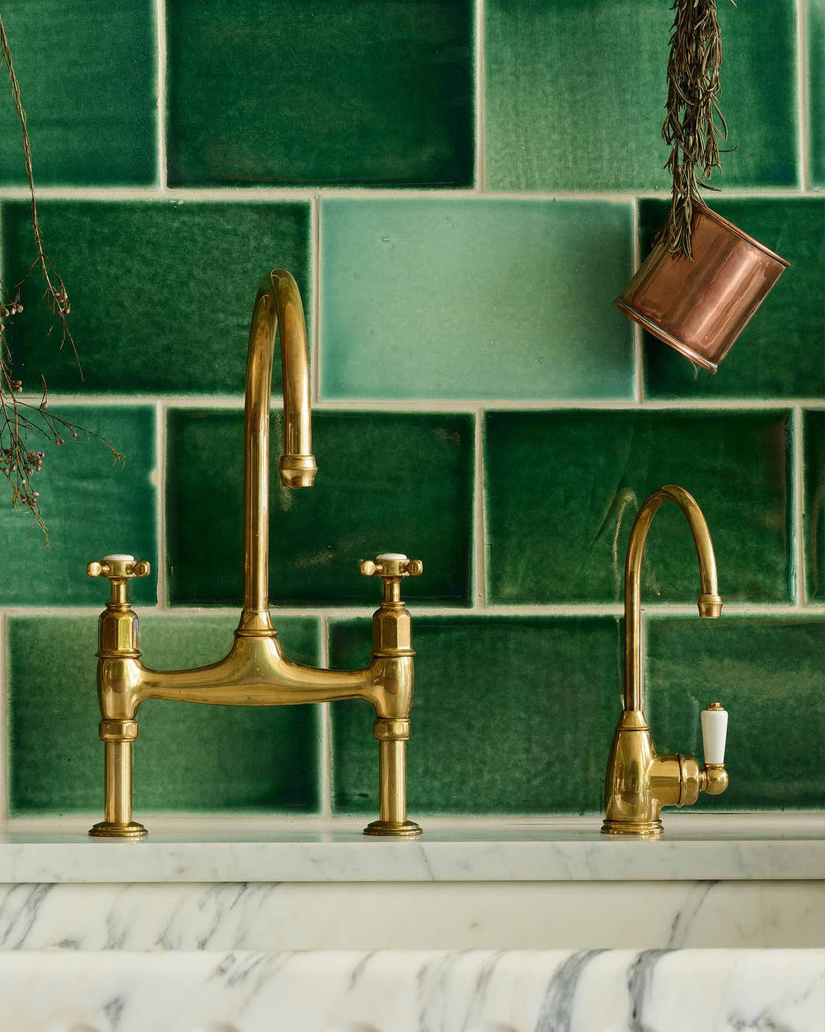 A straight-on view of a small traditional-style aged brass tap beside a larger aged brass mixer tap, with glossy emerald green wall tiles behind. TEST