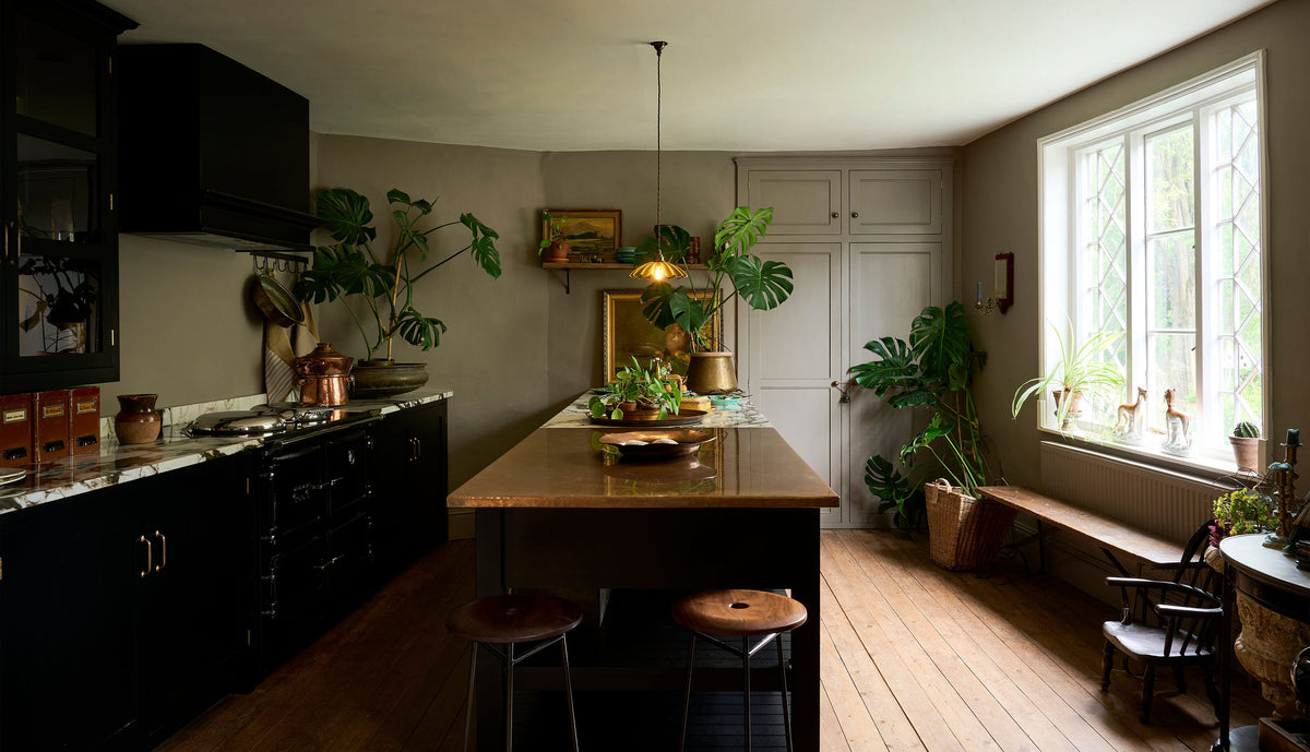 An exotic and glamorous kitchen with black painted Shaker cupboards, warm grey walls with a fitted Classic English pantry, opulent copper and quartzite worktops, original worn floorboards and beautiful leaded windows. The room is brought to life by the green and gold accents of three large monstera plants, a dazzling frilly glass chartreuse pendant light, antique oil paintings and other vintage oddities.