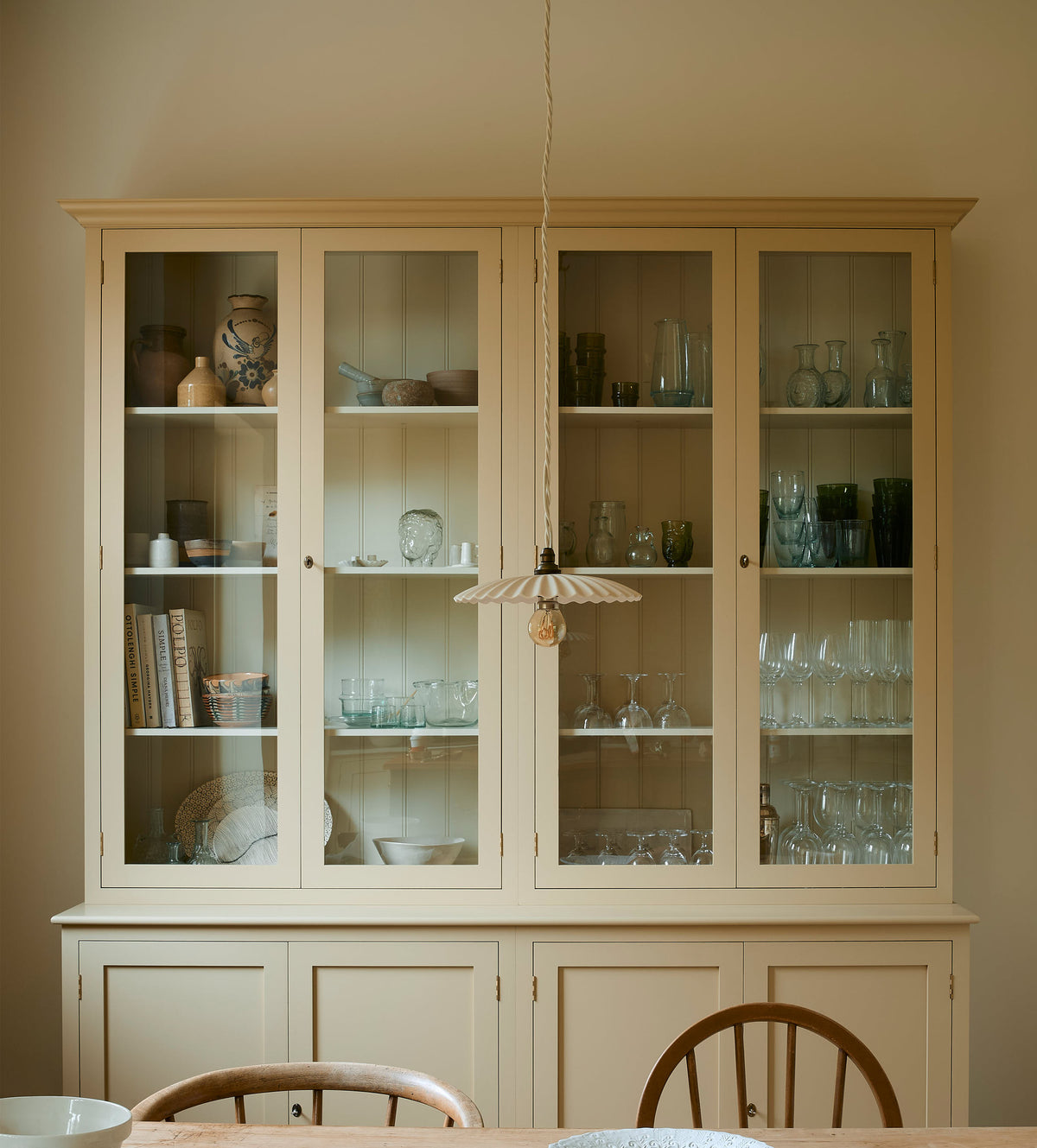  A cream, glazed pantry cupboard stands proudly at the back of a shaker kitchen. In the foreground is an old pine table and antique stick-back chairs, styled with vintage lace, an antique silver spoon and deVOL’s heirloom round platter. A delicate frilly pendant light hangs above the table.