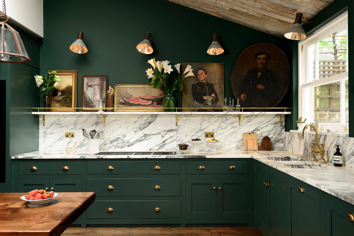 A lush green, classic English kitchen featuring an L-shaped sink and cooker, all complemented by Arabescato marble for the worktop, splashback, and countertop shelf. The kitchen includes full-height cabinets and a wall cupboard, creating a luxurious yet bohemian atmosphere. An angled wooden skylight window allows natural light to flow into the room, highlighting delicate bird-painted plates, a rich oak dining table, framed antique paintings, and industrial-style gas light fixtures on the walls.