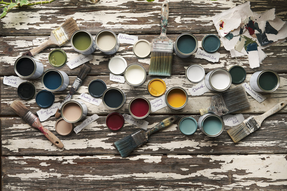 An overhead view of open paint pots with a variety of different colours on a worn painted tabletop.