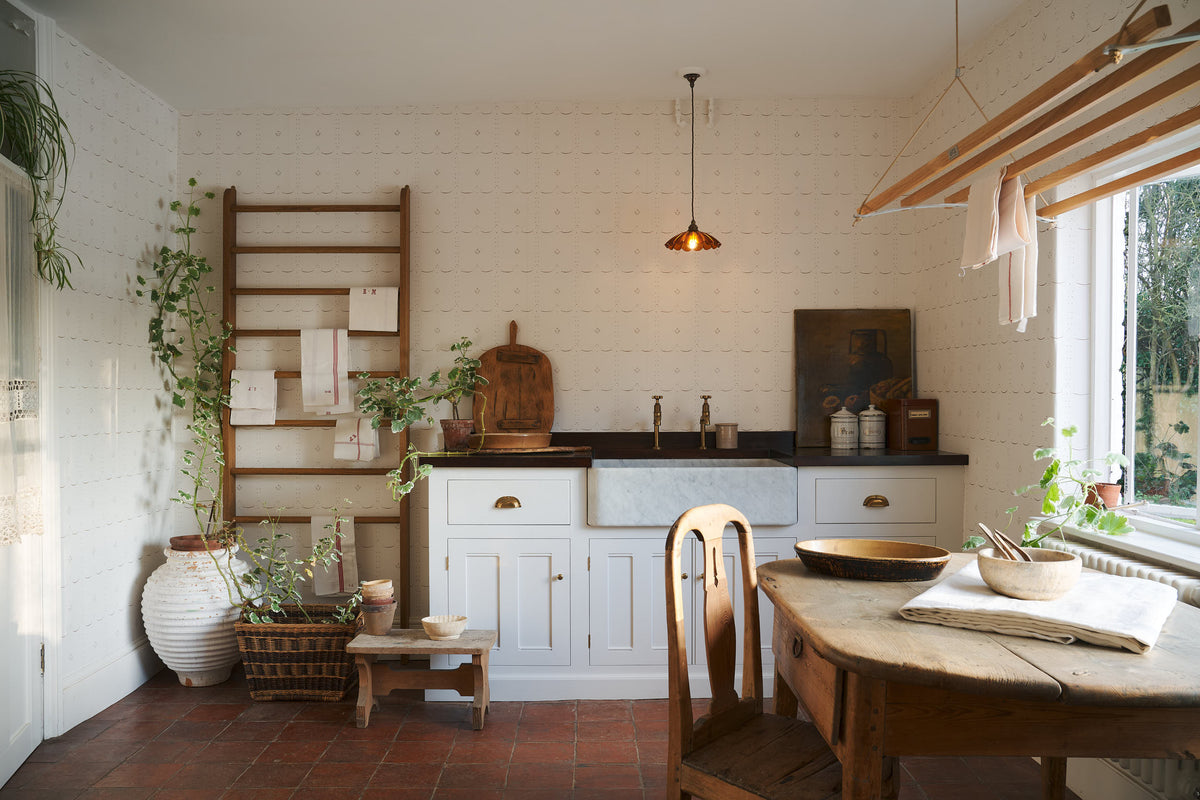 A Mediterranean-inspired scullery with a terracotta square floor, linen white sink run, a frilly glass amber light, a laundry maid and a delicate off-white sepia ditsy delft wallpaper.
