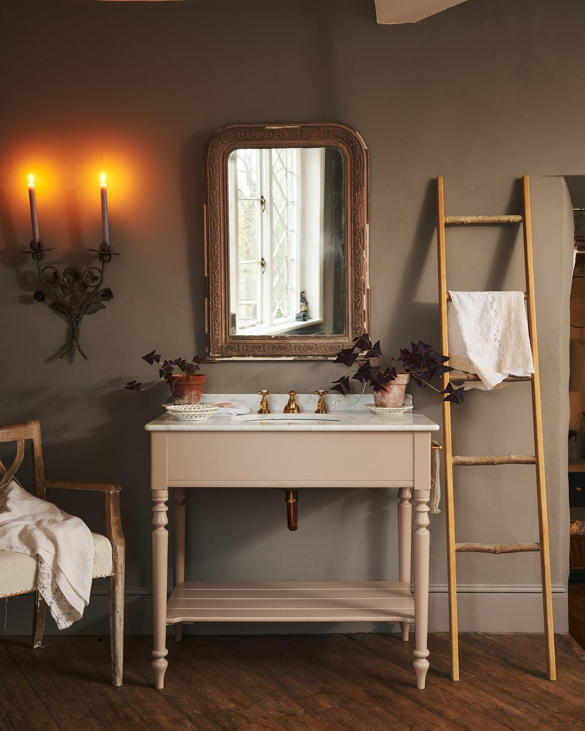 An oblique view of a dusty-pink Victorian-inspired washstand with brass mixer tap in a greyish-brown painted room. The shot is atmospheric and moody, with two lit candles casting a subtle glow and light from an adjacent window illuminating the brass accents. | Large Princelet Pink shown TEST