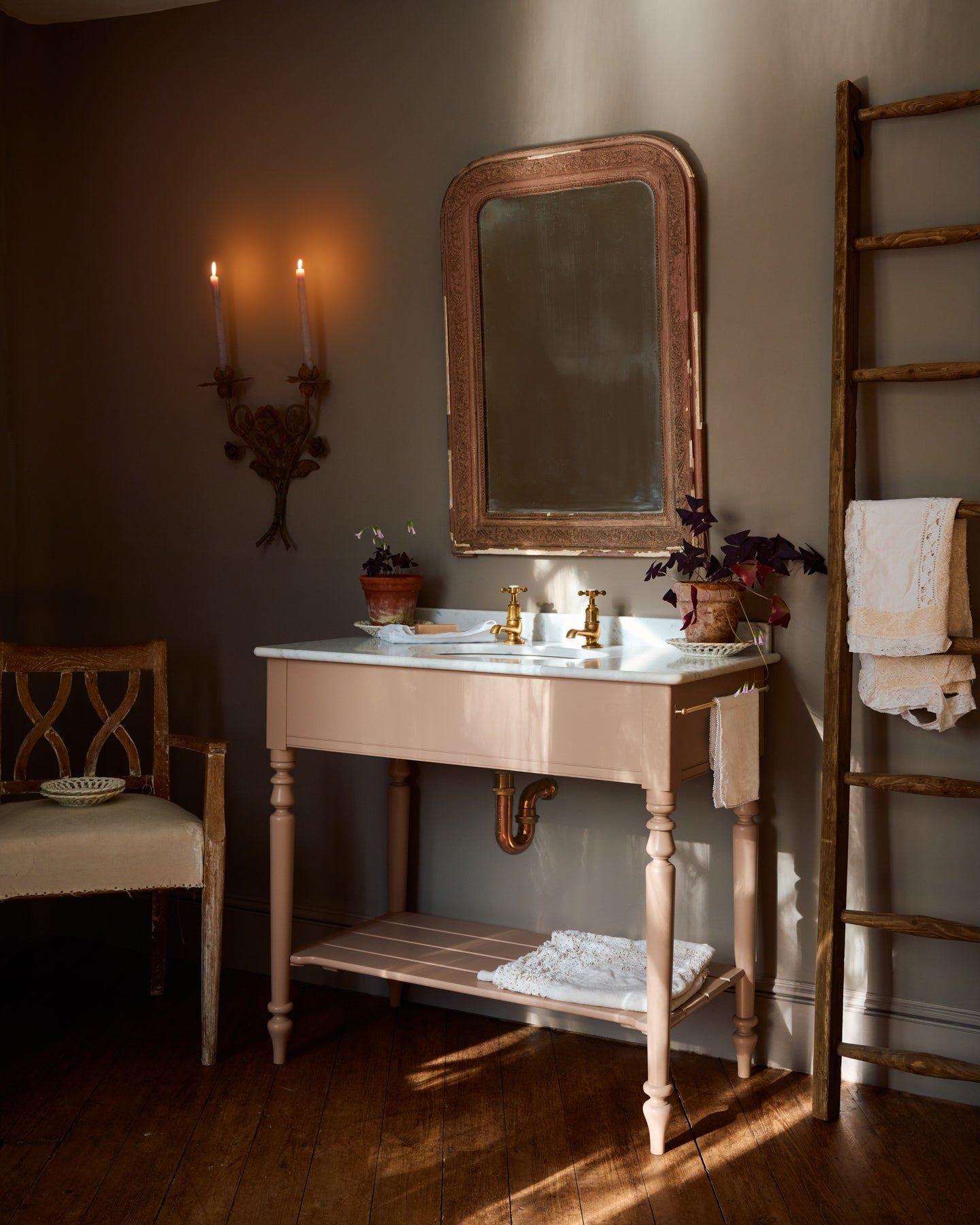 An oblique view of a dusty-pink Victorian-inspired washstand with brass pillar taps in a greyish-brown painted room. The shot is atmospheric and moody, with two lit candles casting a subtle glow and light from an adjacent window illuminating the brass accents.