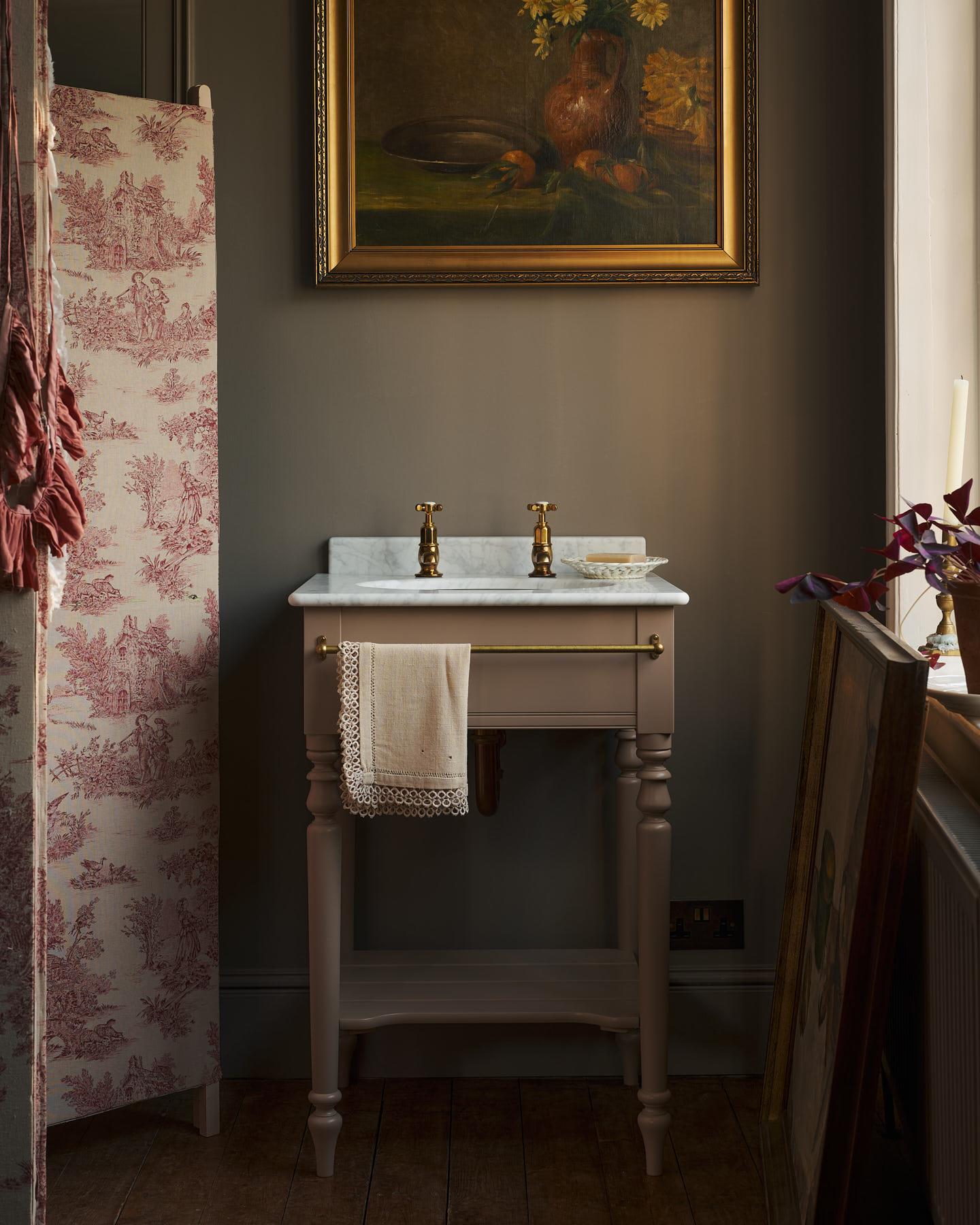 A straight-on view of a mushroom-grey Victorian-inspired washstand with a brass mixer tap in a duck-egg blue painted room. The room is classic and bohemian with an ornate white mirror and elegant painted flowers across the top of the walls.