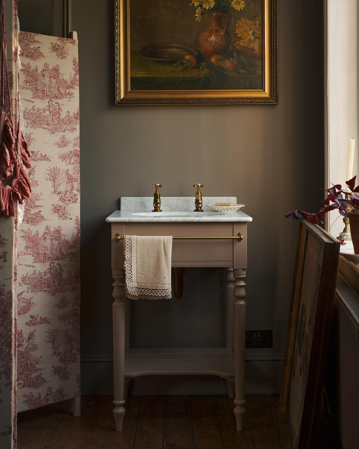 A straight-on view of a mushroom-grey Victorian-inspired washstand with a brass mixer tap in a duck-egg blue painted room. The room is classic and bohemian with an ornate white mirror and elegant painted flowers across the top of the walls. | Princelet Pink with Pillar Taps shown TEST