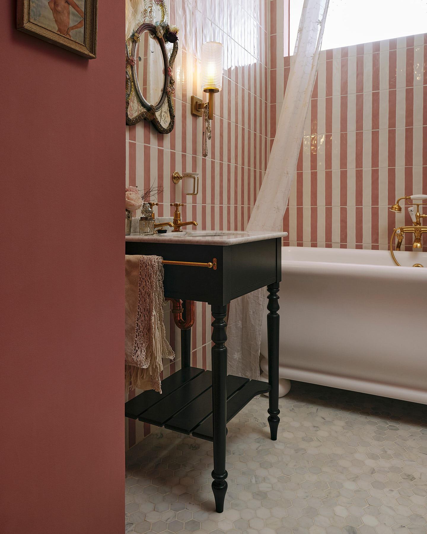 An oblique view of a porcelain bath and a black Victorian-inspired washstand with a brass pillar tap against a pink and white splashback tiles. A decorative antique mirror sits above the sink, along with deco-style scones, and a brass cup holder on the wall.