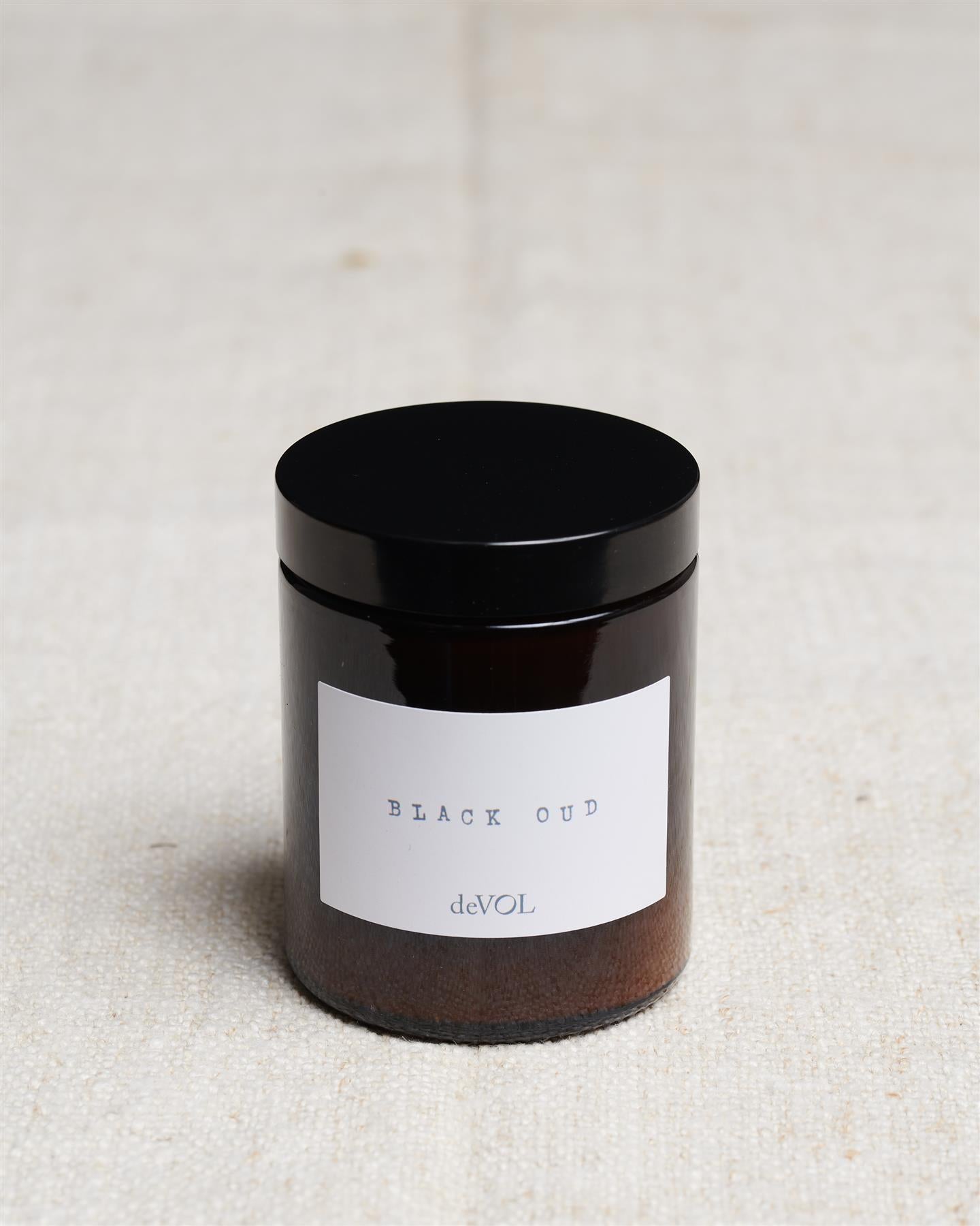 A medium scented candle in a brown glass jar with a black lid, the label reads 'Amber & Tonka Bean, deVOL'.