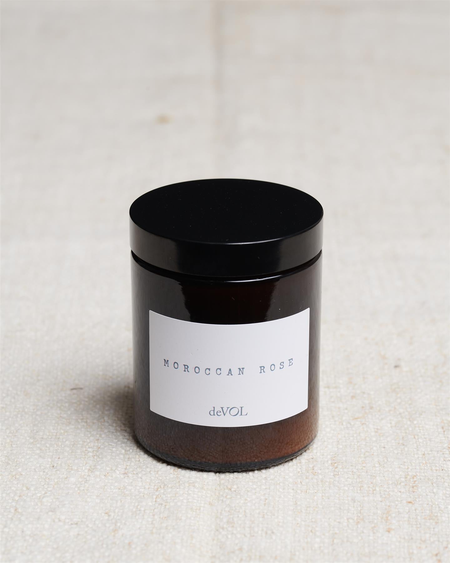 A medium scented candle in a brown glass jar with a black lid, the label reads 'Amber & Tonka Bean, deVOL'.