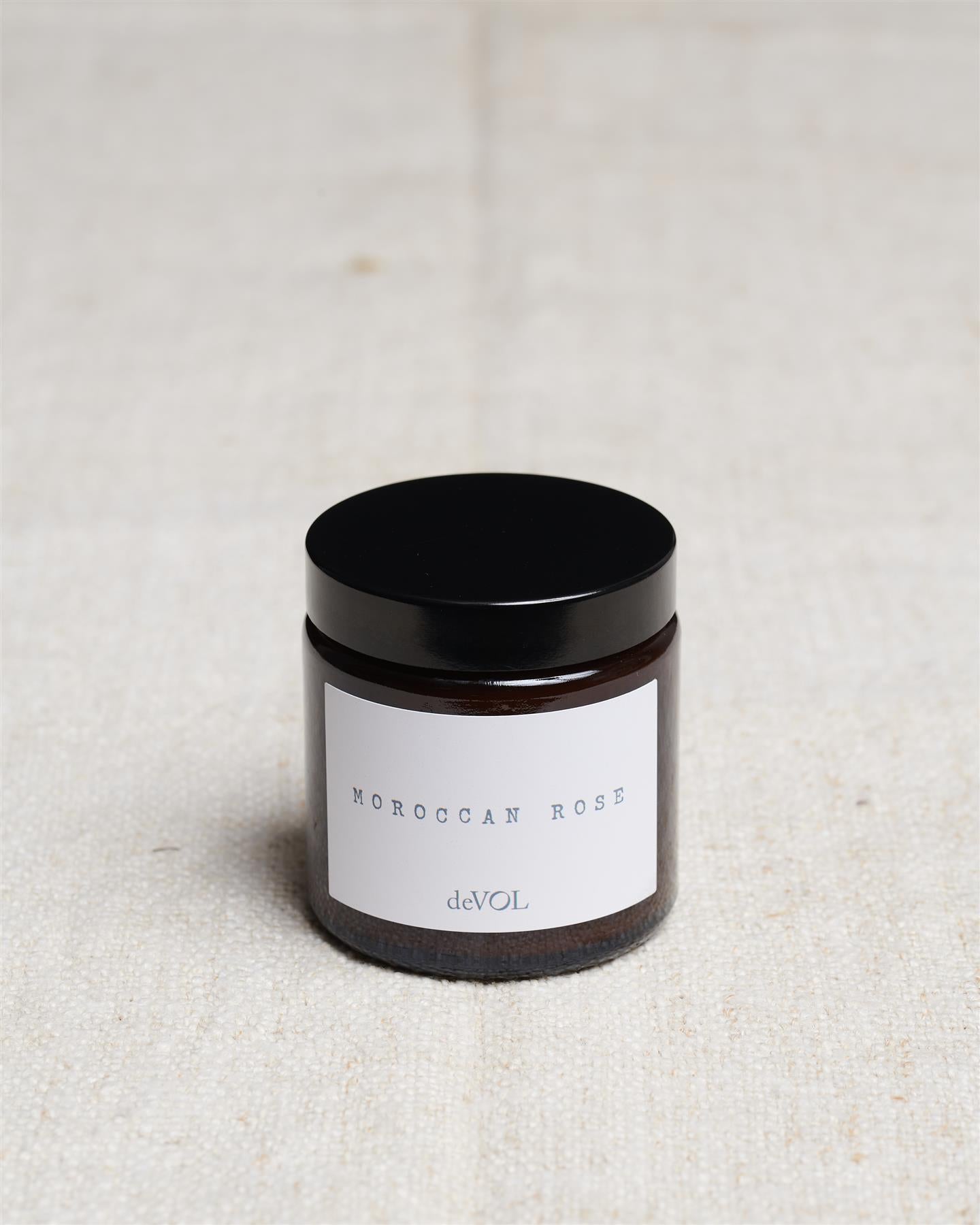 A small scented candle in a brown glass jar with a black lid, the label reads 'Amber & Tonka Bean, deVOL'.