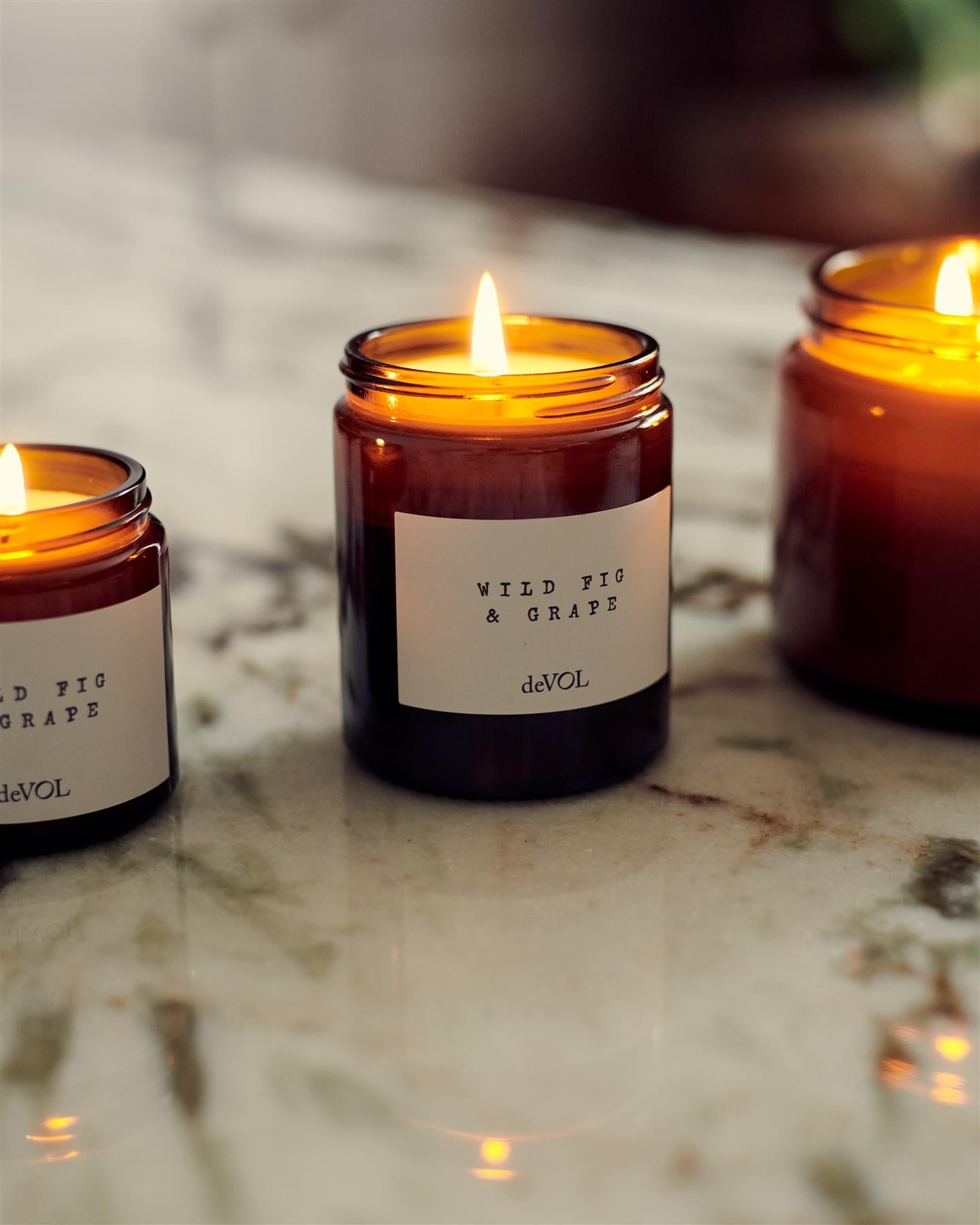 A straight-on view of a collection of three lit scented candles in brown glass jars on a wooden stool top; small, medium and large.