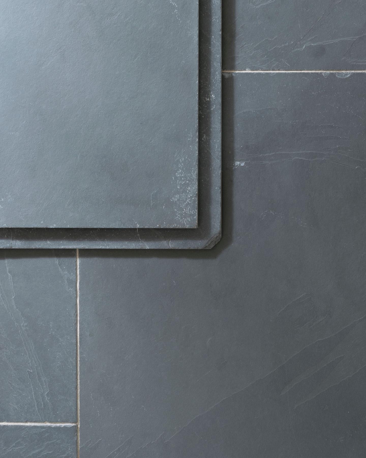 A close-up oblique view of a grey tumbled slate floor, with two loose tiles stacked on top.