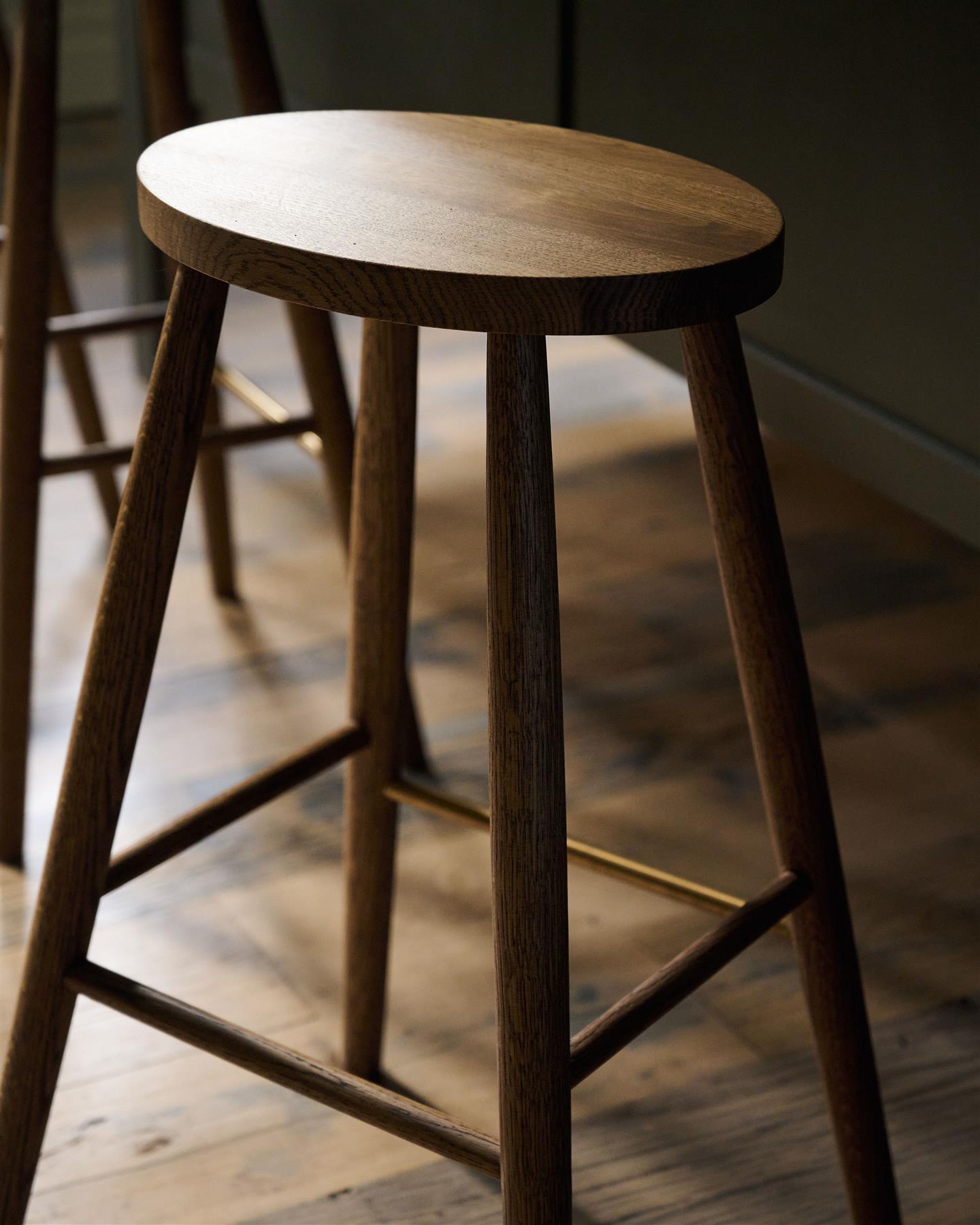 An oblique view of a simple oak bar stool with an oval-shaped seat and a brass footrail.