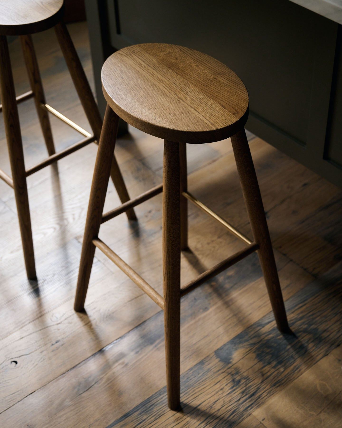 An oblique view of three simple oak bar stools with oval-shaped seats in front of a dark green painted Shaker-style island breakfast bar.