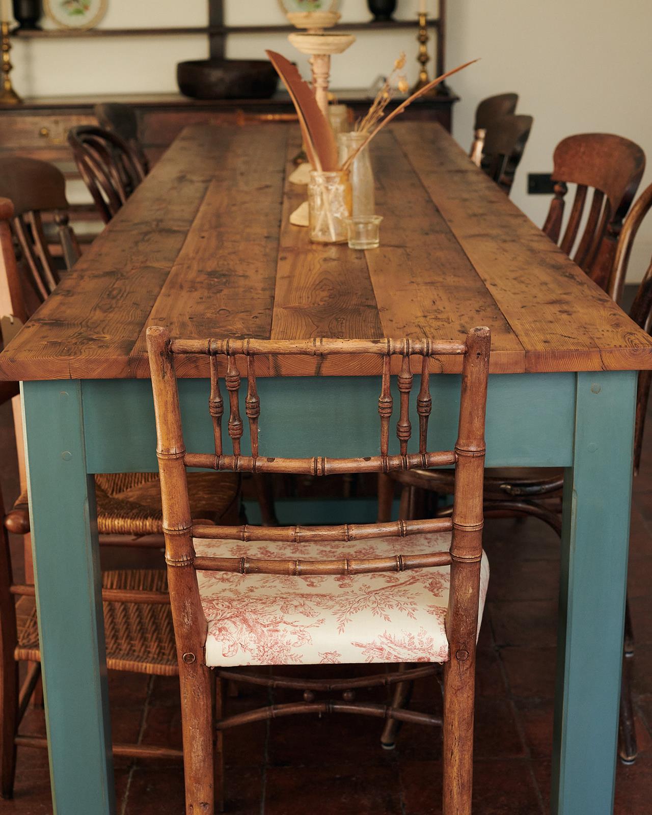 An antique wooden chair at the end of a long banquet-style dining table with simple blue-green painted legs and frame and rustic reclaimed pine boards for the tabletop.