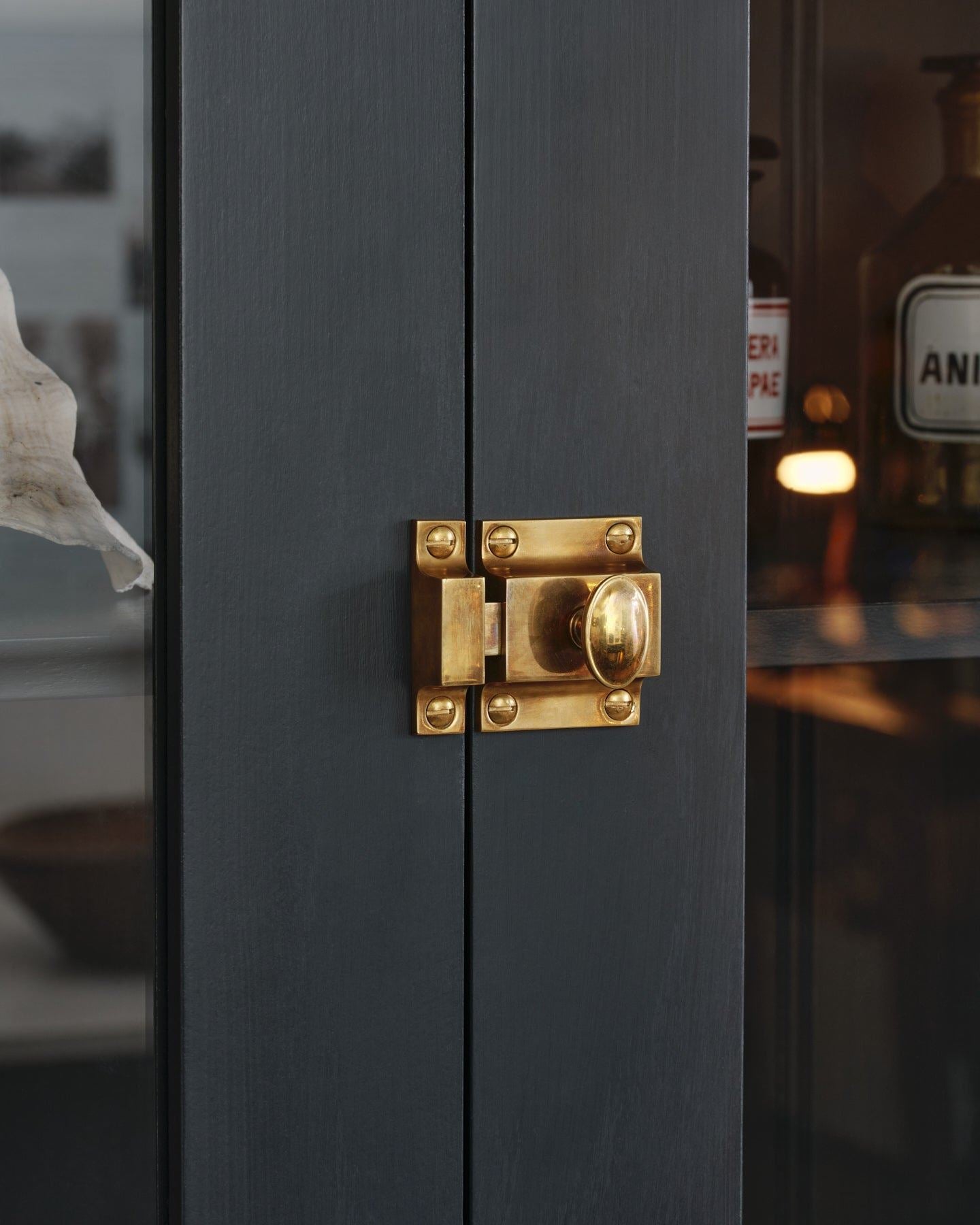 An oblique view of a classic-style square cupboard catch with an ornate oval-shaped knob in a warm burnished brass finish fitted to dark blue painted glazed cupboard doors.