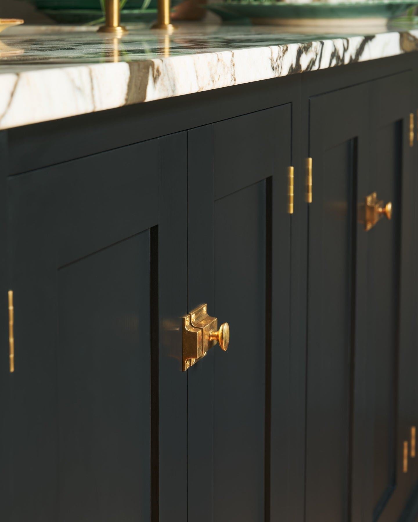 An oblique view of classic-style square cupboard catches in a warm burnished brass finish fitted to black painted kitchen cupboard doors.