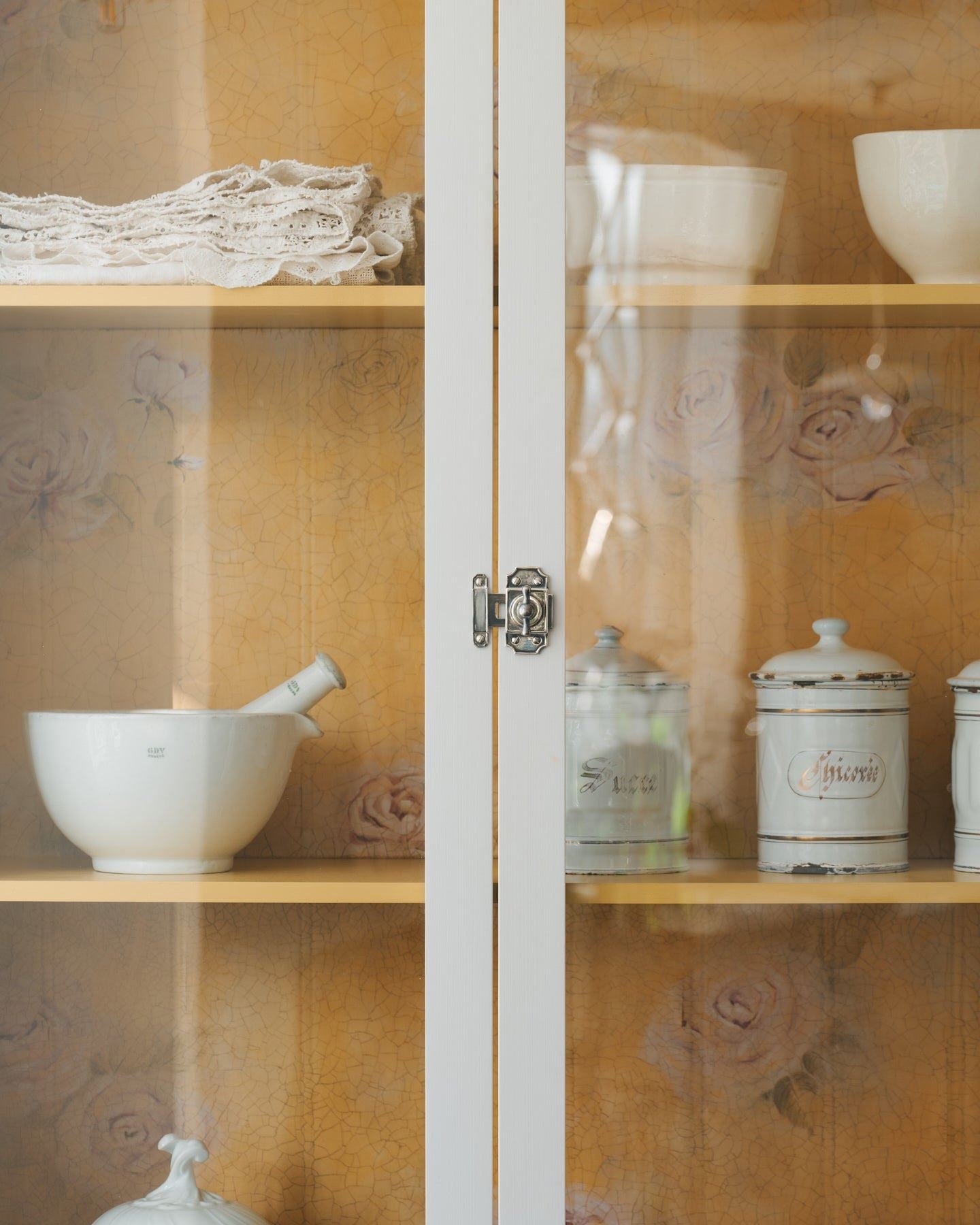 A straight-on view of a Victorian-inspired rack and pinion cupboard catch in a tarnished silver finish, fitted to a tall glazed cupboard with white painted doors and a yellow painted interior.
