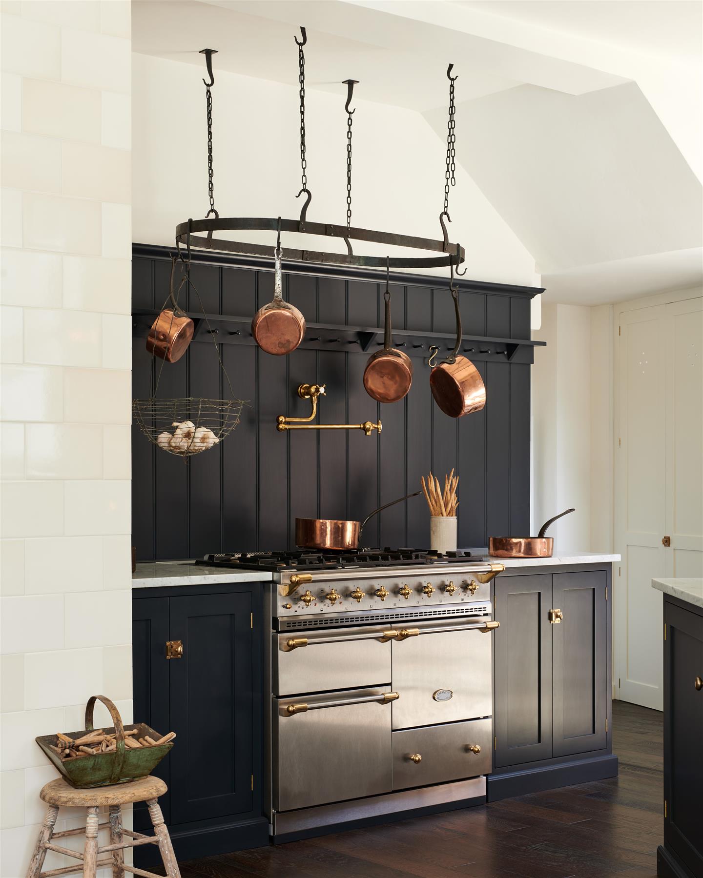 An oblique view of a simple Shaker-style kitchen with dark blue painted cabinetry and a stainless steel Lacanche range cooker with a large oval-shaped wrought iron pot hanger suspended from chains above.