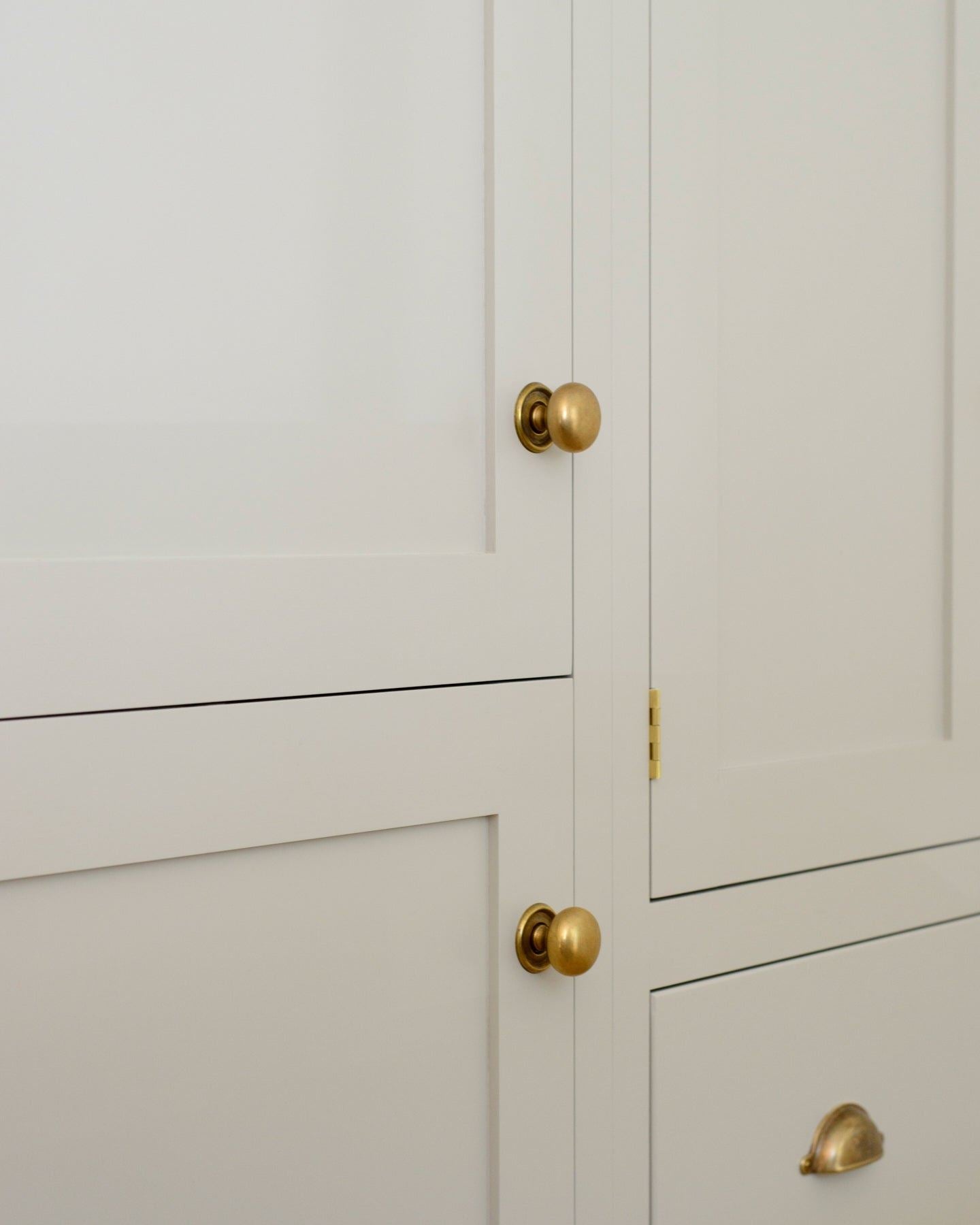 An oblique view of a cream painted Shaker-style pantry cupboard with a set of round doorknobs in a burnished 'Bella' brass finish.