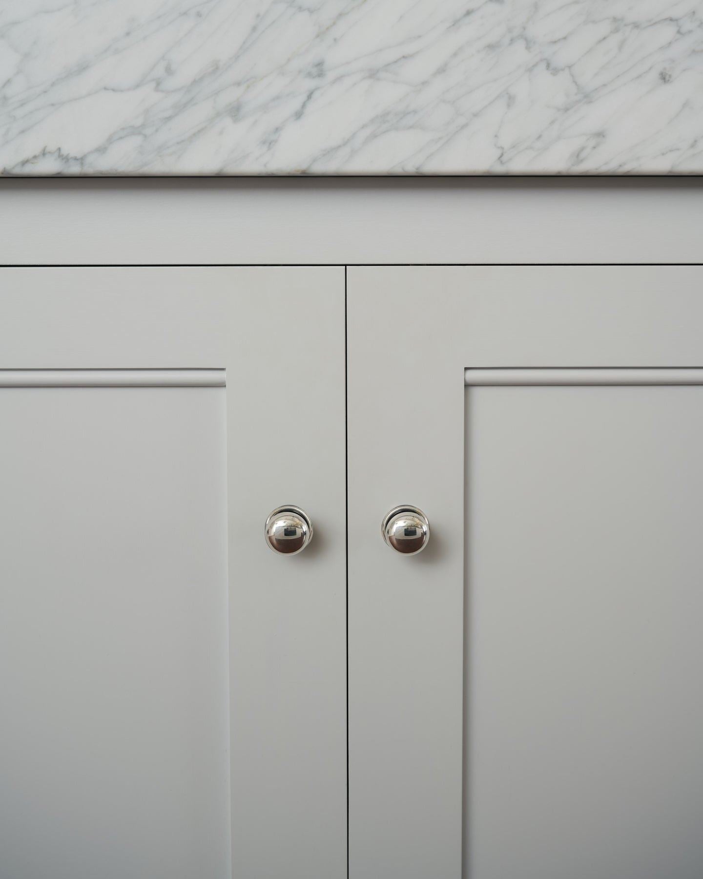 A straight-on view of a pair of round doorknobs with pressed backplates in a tarnished silver finish, fitted to white painted Georgian-style kitchen cupboard doors.