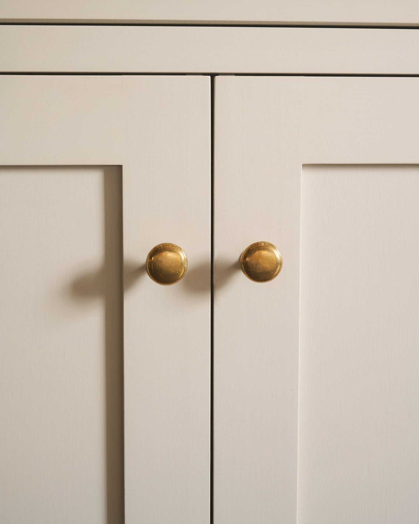 A straight-on view of a pair of classic knobs in a warm burnished brass finish, fitted to a mushroom painted Shaker-style kitchen cupboard.