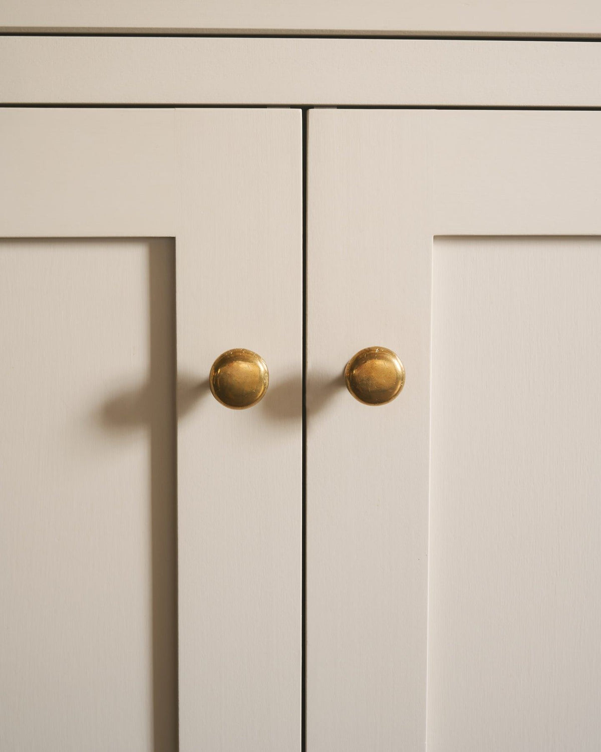 A straight-on view of a pair of classic knobs in a warm burnished brass finish, fitted to a mushroom painted Shaker-style kitchen cupboard.|Heirloom Brass Medium shown TEST