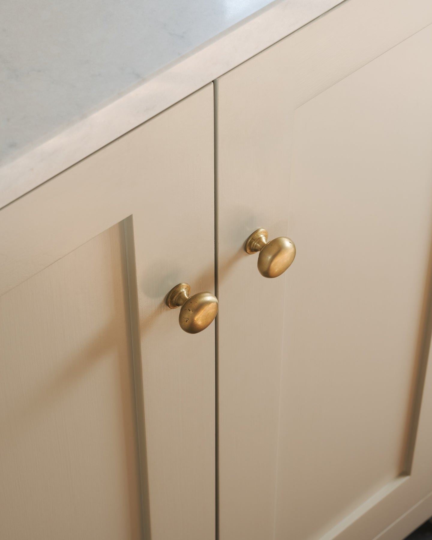 An oblique view of a pair of classic knobs in an aged brass finish, fitted to a mushroom painted Shaker-style kitchen cupboard.