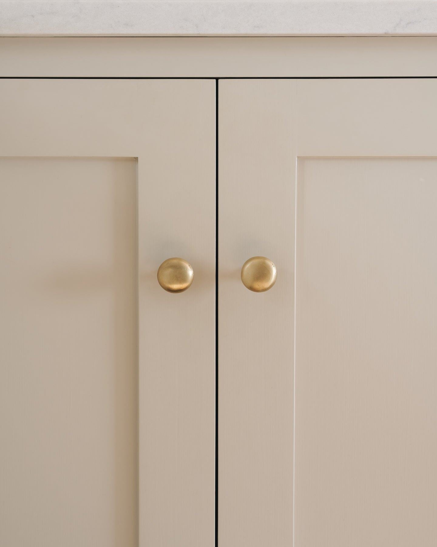 A straight-on view of a pair of classic knobs in an aged brass finish, fitted to a mushroom painted Shaker-style kitchen cupboard.