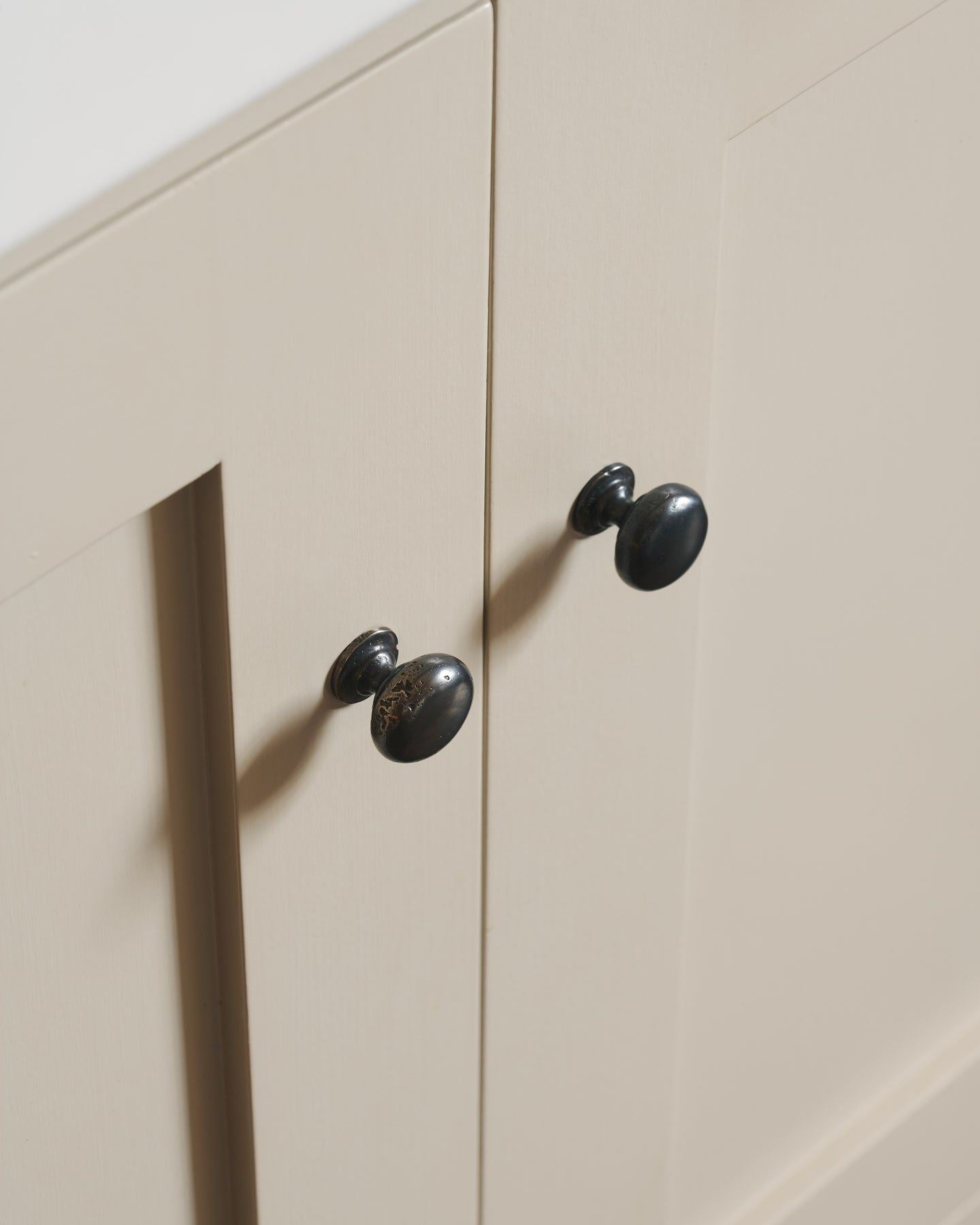 An oblique view of a pair of classic knobs in a dark oxidised bronze finish, fitted to a mushroom painted Shaker-style kitchen cupboard.