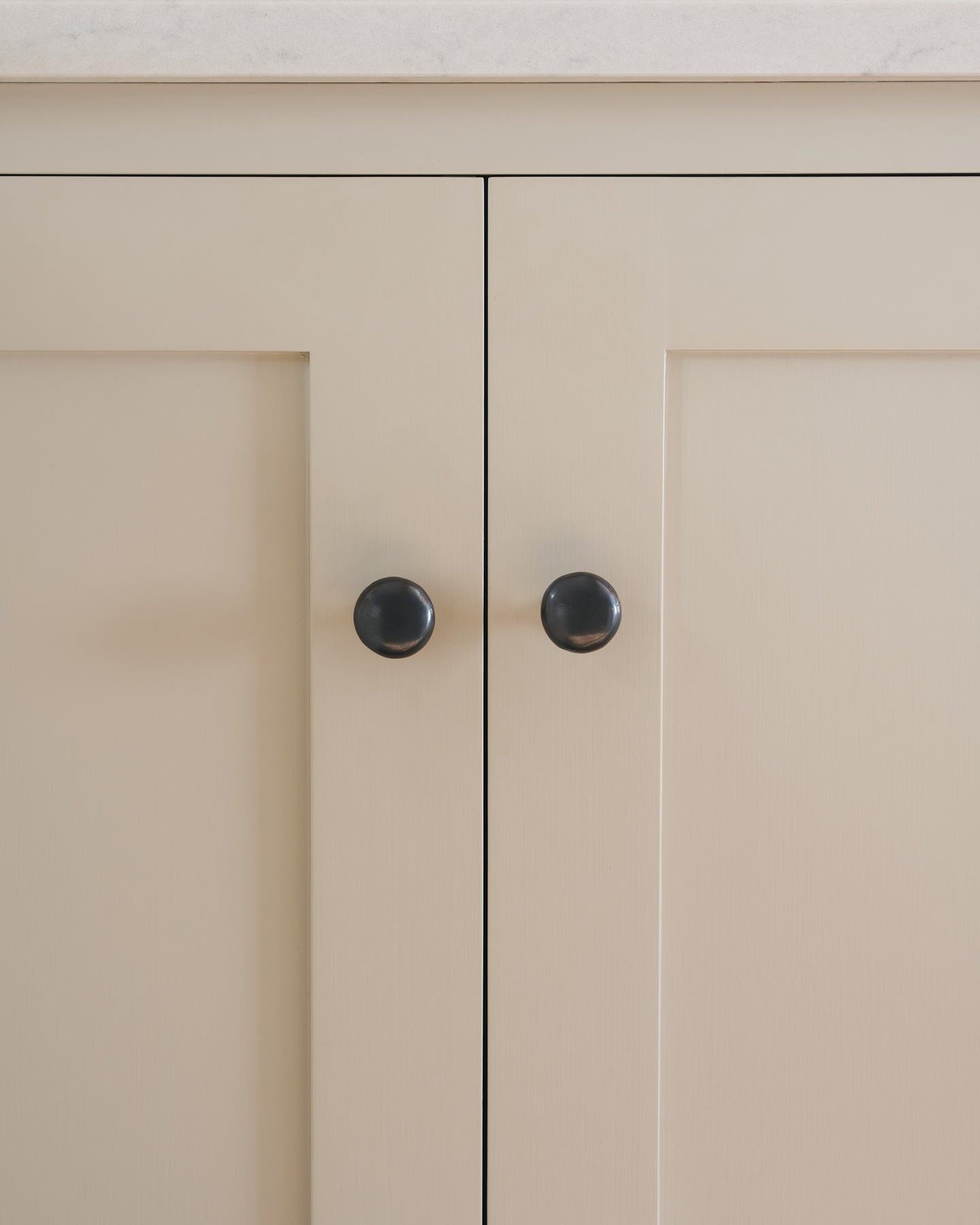 A straight-on view of a pair of classic knobs in a dark oxidised bronze finish, fitted to a mushroom painted Shaker-style kitchen cupboard.