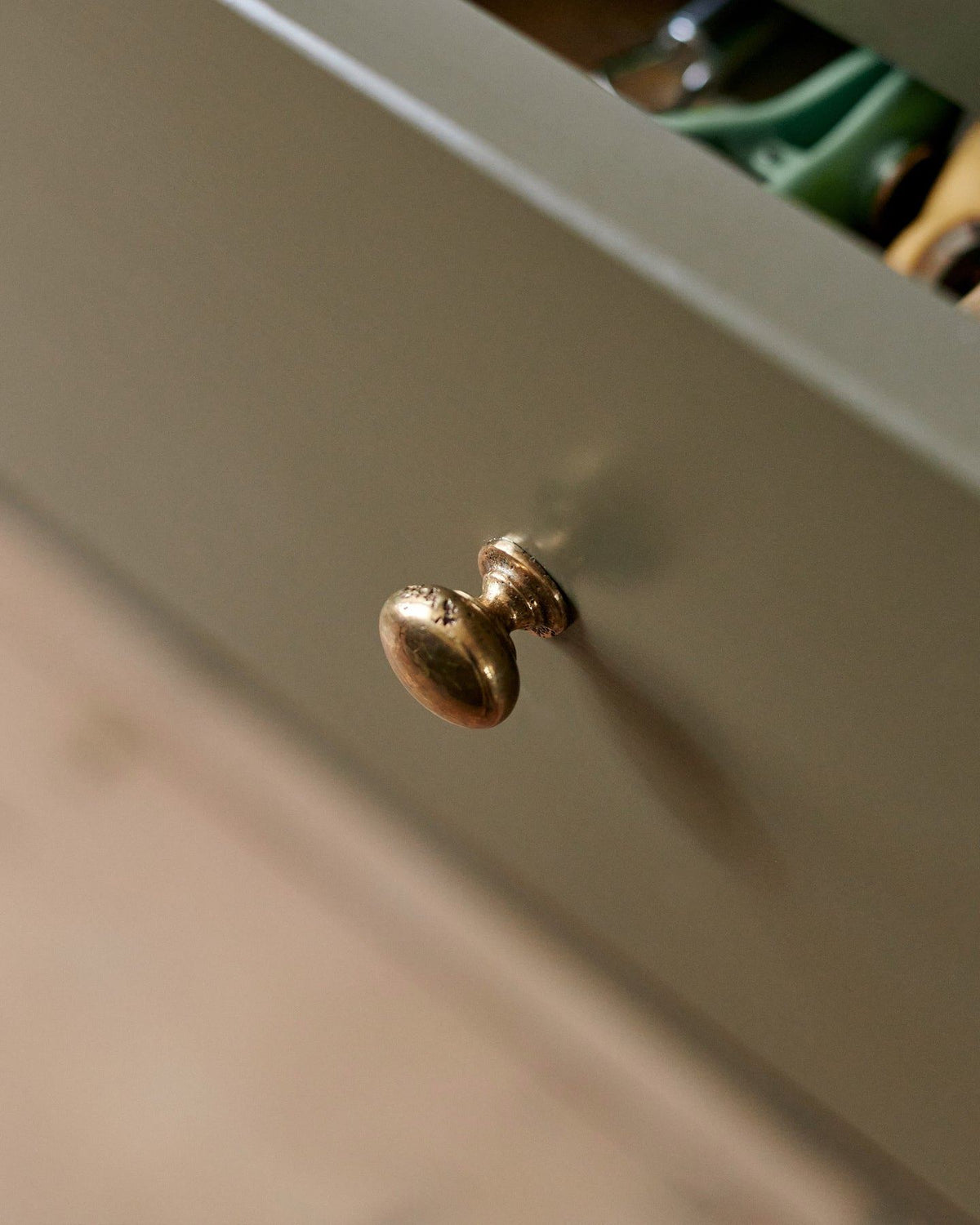 A close-up oblique view of a classic knob in a bright polished bronze finish, fitted to a mossy green painted drawer front. TEST