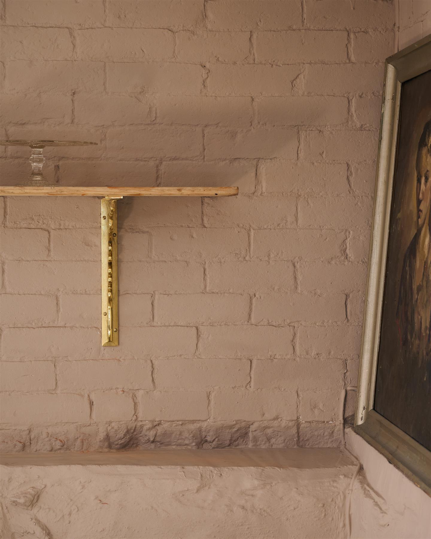 A straight-on view of a large Victorian-inspired ornate aged brass shelf bracket holding a deep pine shelf to a pink colour brick wall.
