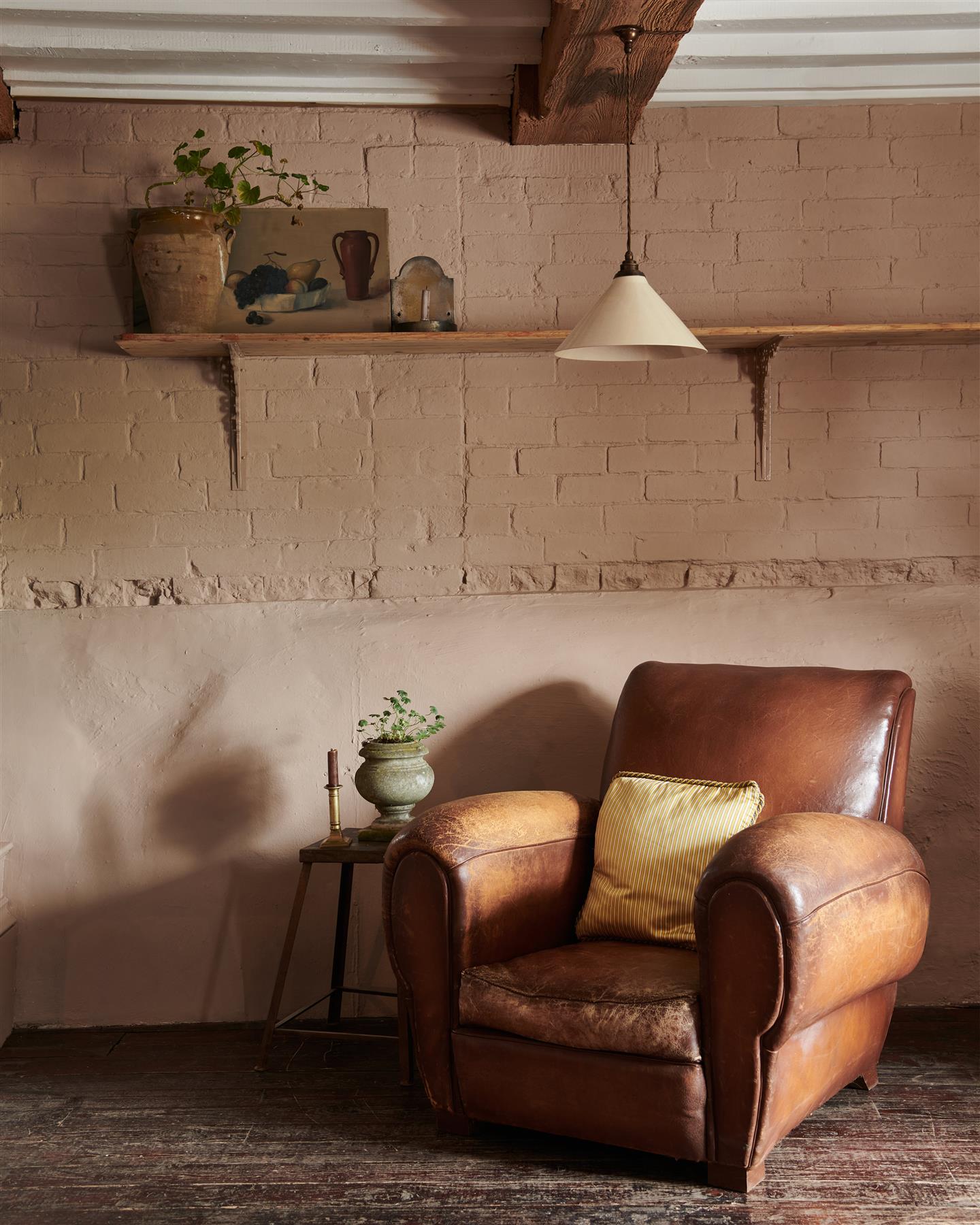 A vintage leather armchair in front of a pink brick wall and long pine shelf with Victorian-inspired ornate pink painted shelf brackets.