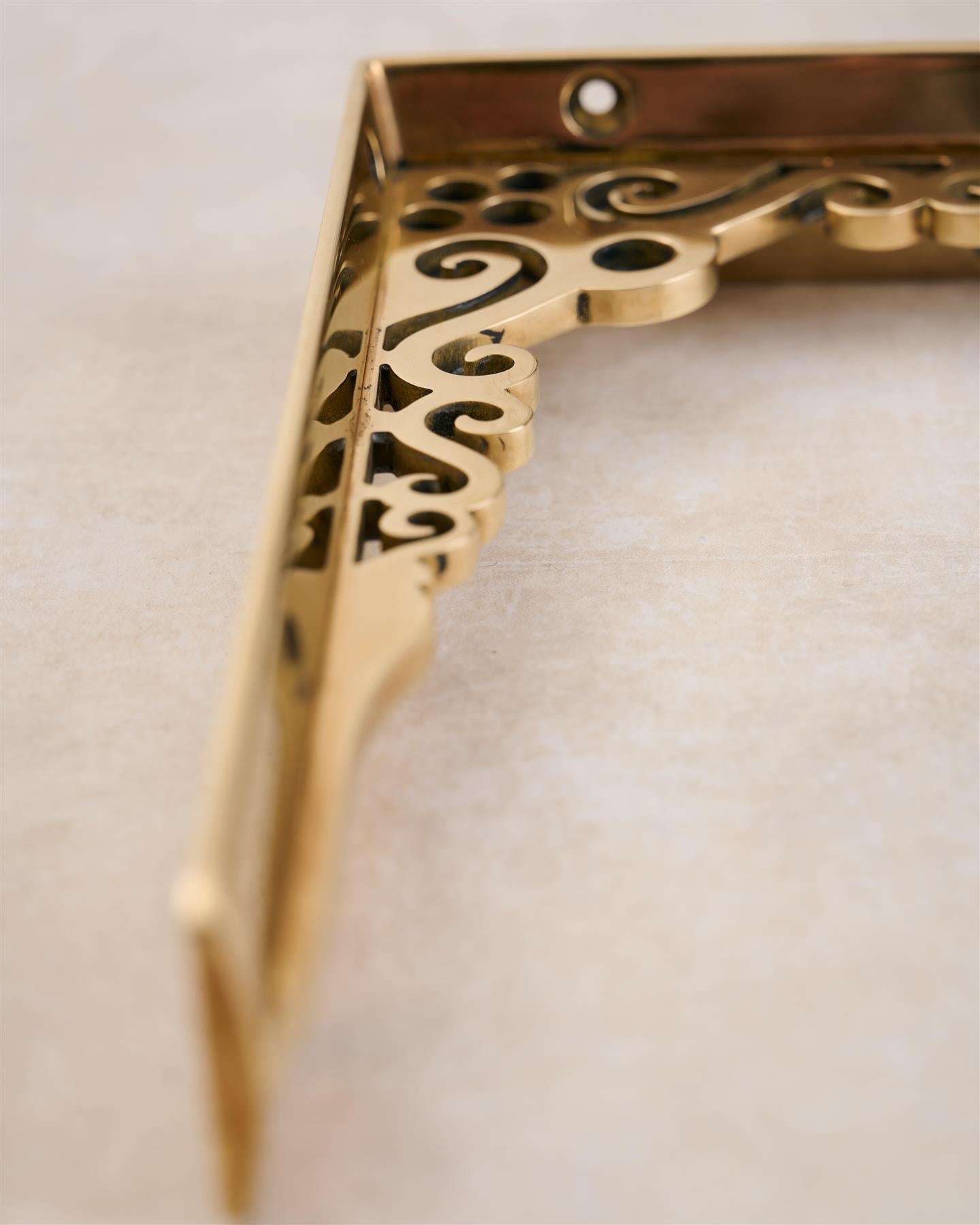 A close-up view of the decorative cutout patterns on a small Victorian-inspired ornate polished brass shelf bracket.