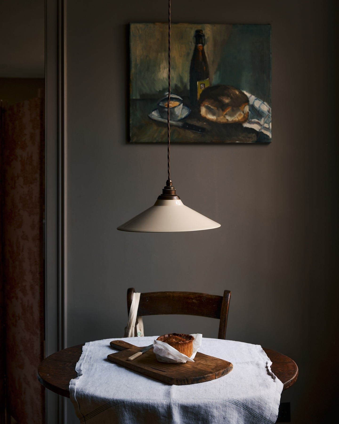 A straight-on view of a rise and fall light with a small cream glazed ceramic shade, aged brass fittings and brown twisted fabric flex cord hanging in a dark room over a small round wooden table with a linen tablecloth and fresh bread.