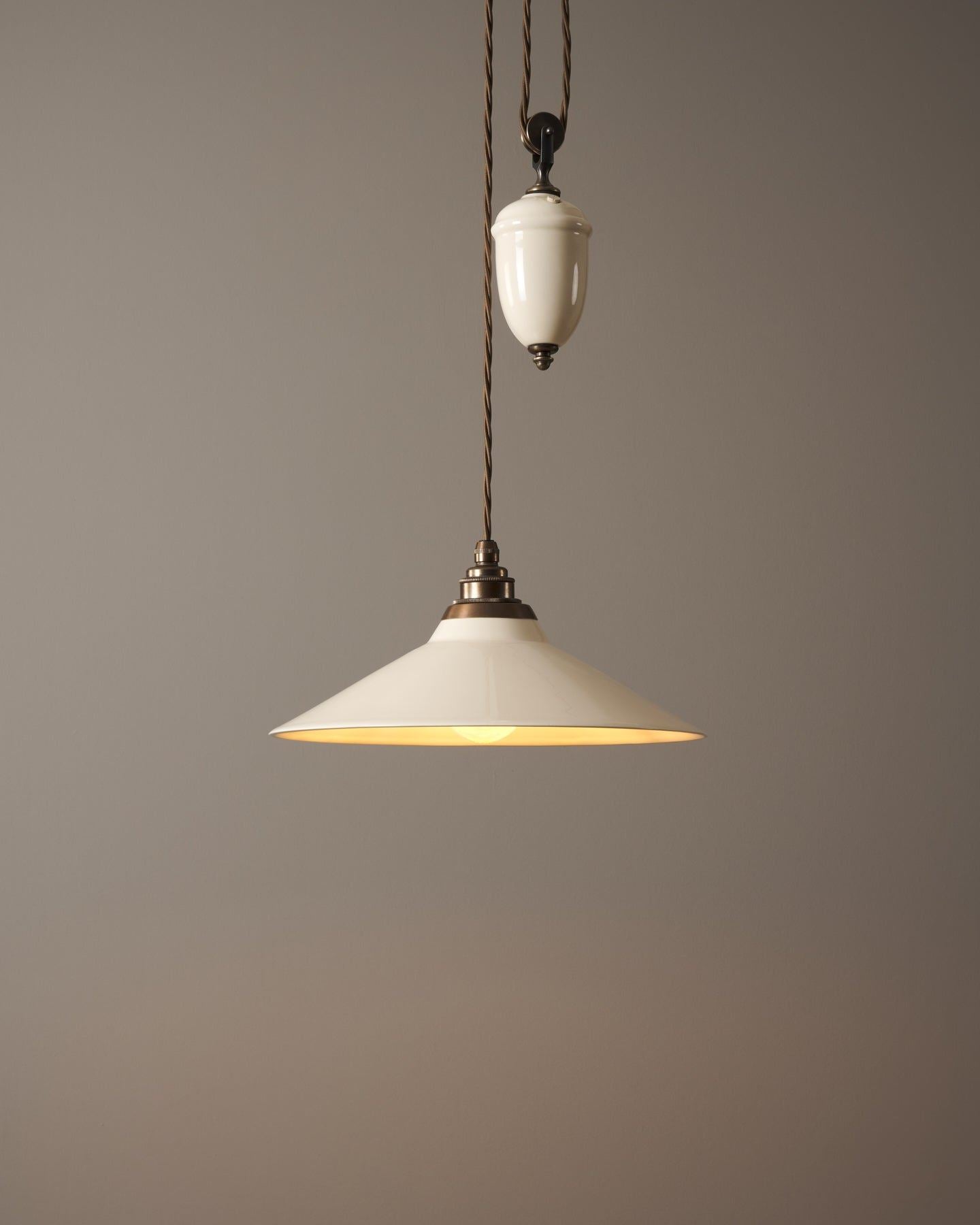 A rise and fall light with a small cream glazed ceramic shade and counterweight, an aged brass E27 lampholder and brown twisted fabric flex cord, the light is turned on.