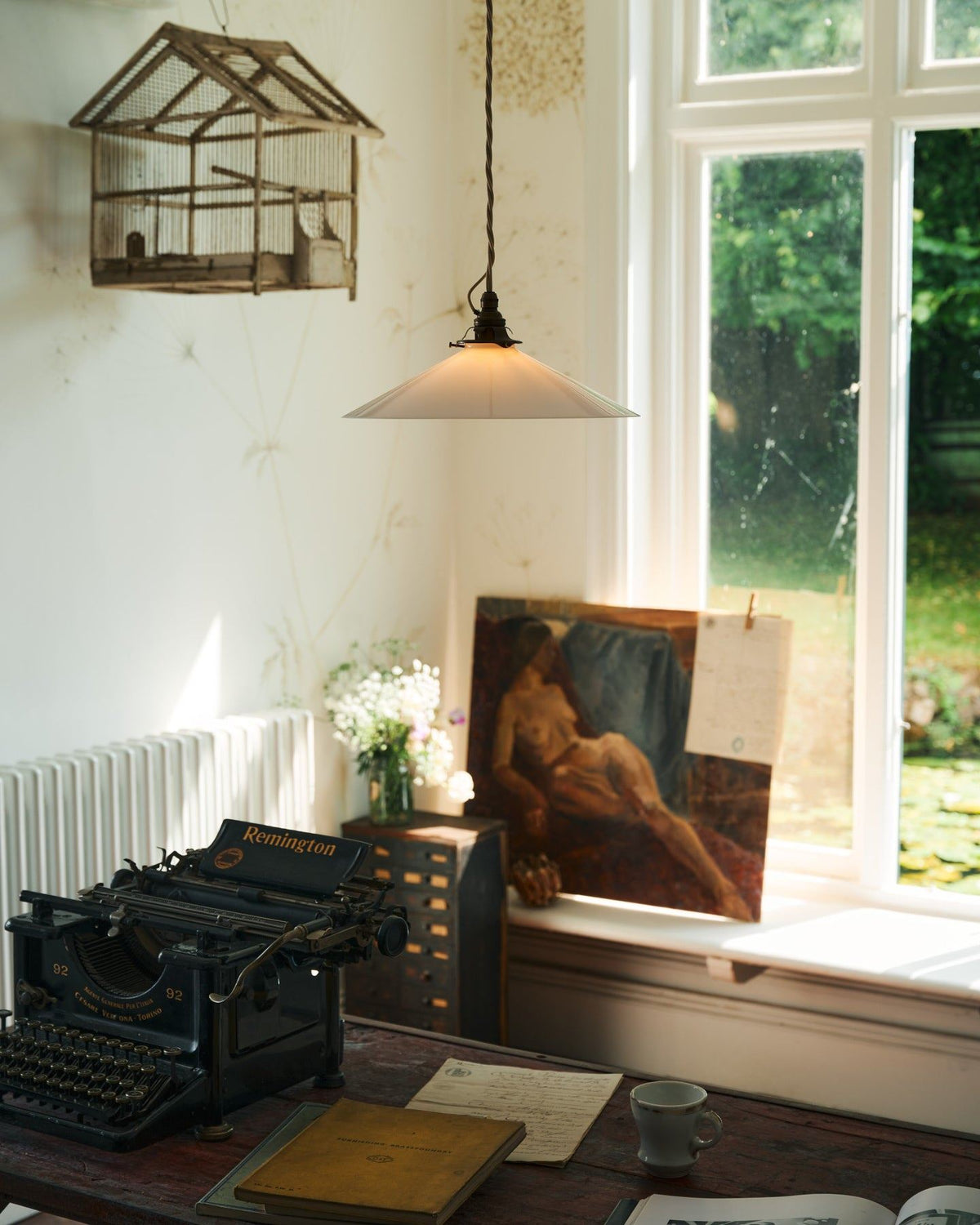 An oblique view of a classic pendant light with a milk glass light shade, brass fittings and a cream twisted fabric flex cord, hanging above a typewriter and scraps of paper by an old oil painting and a large window. TEST