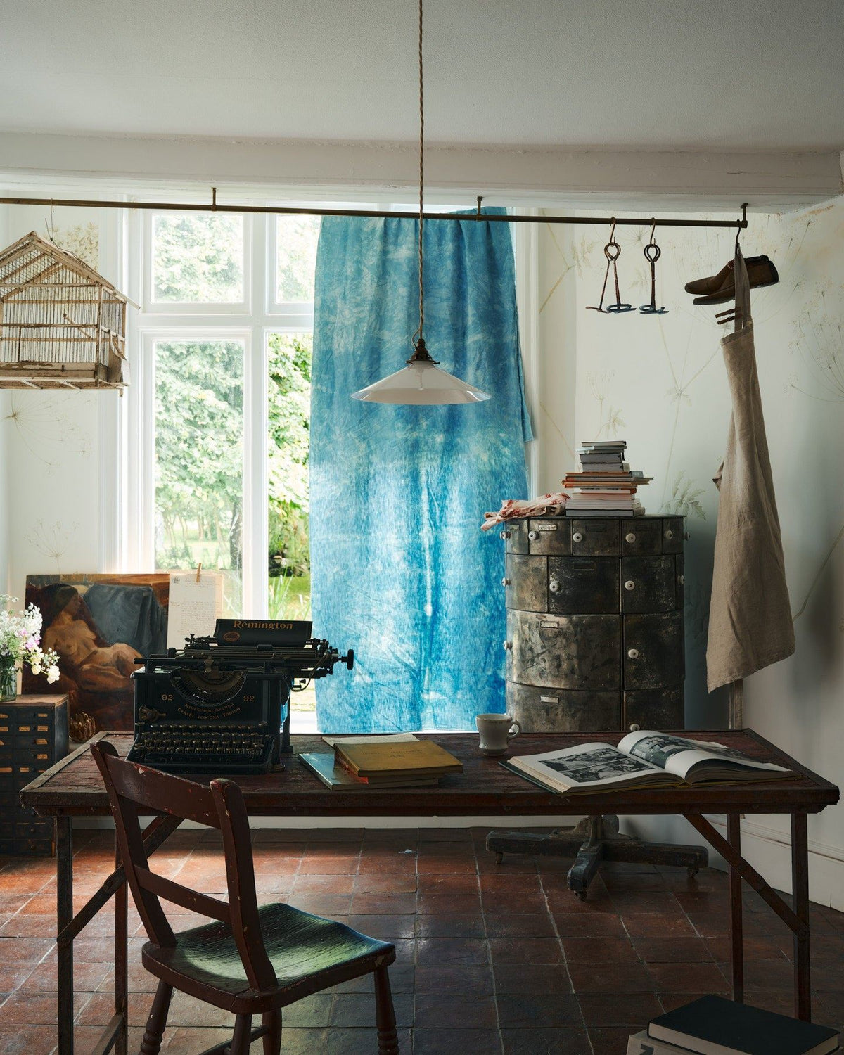 A straight-on view of a classic pendant light with a milk glass light shade, brass fittings and a cream twisted fabric flex cord, hanging above a typewriter and scraps of paper. TEST
