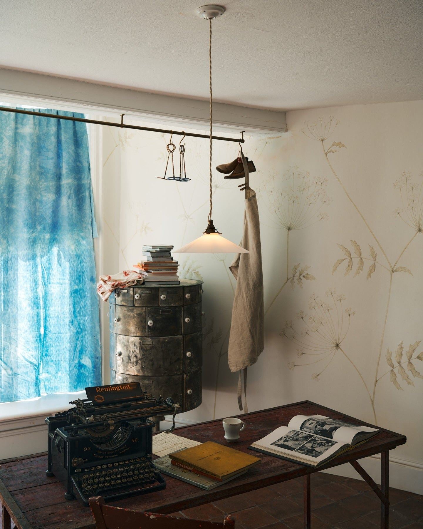 An oblique view of a classic pendant light with a milk glass light shade, brass fittings and a cream twisted fabric flex cord, hanging above a typewriter and scraps of paper.