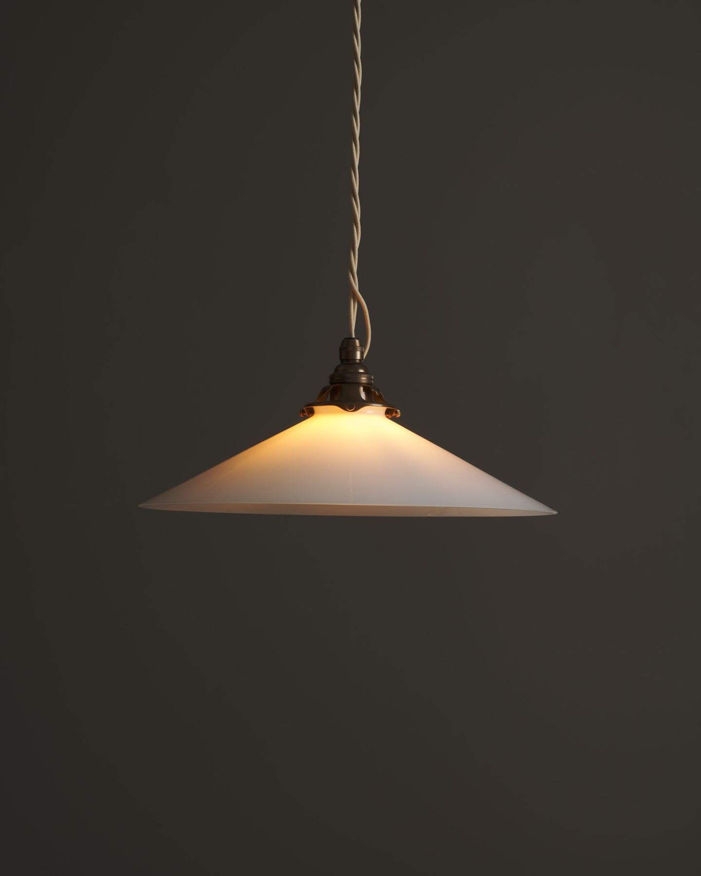 A classic pendant light with a milk glass light shade, an aged brass E27 lampholder with a decorative gallery and cream twisted fabric flex cord. The light is on, showing the warm red glow of the translucent surface.
