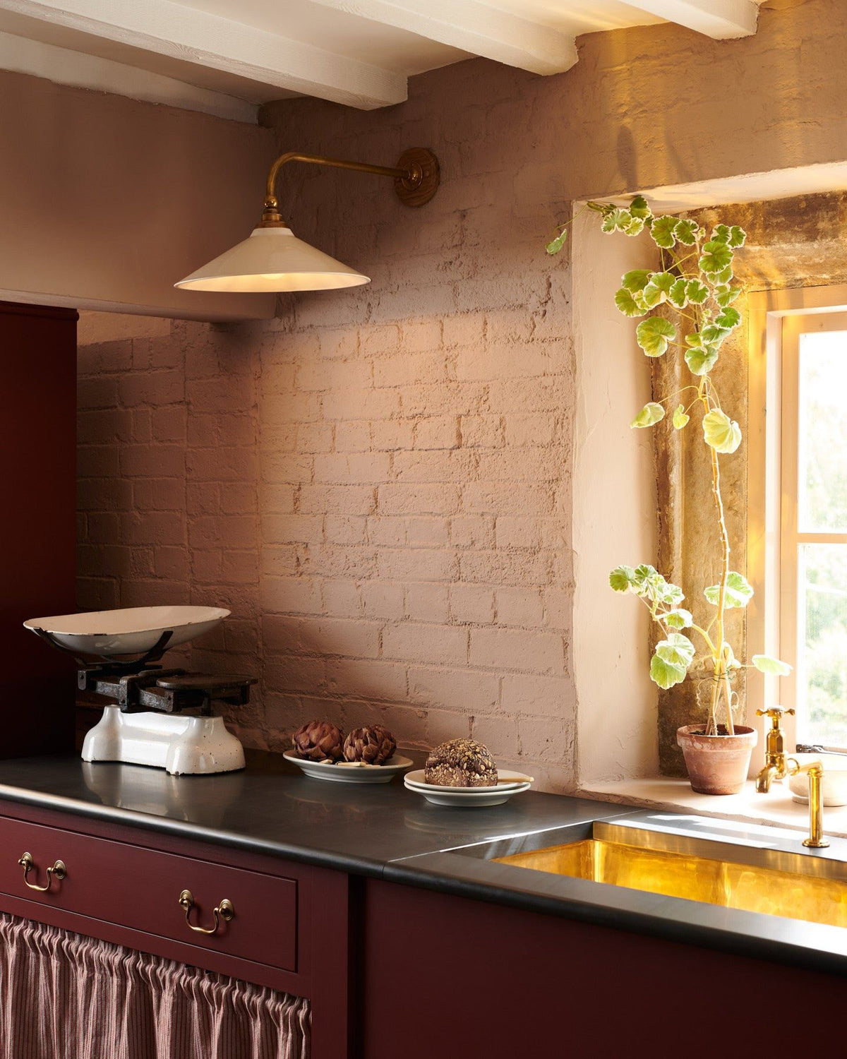 An oblique view of a large Victorian-inspired wall light with a brass arm and a cream glazed ceramic shade fixed to a pink painted brick wall above a slate worktop and red painted cabinetry. TEST