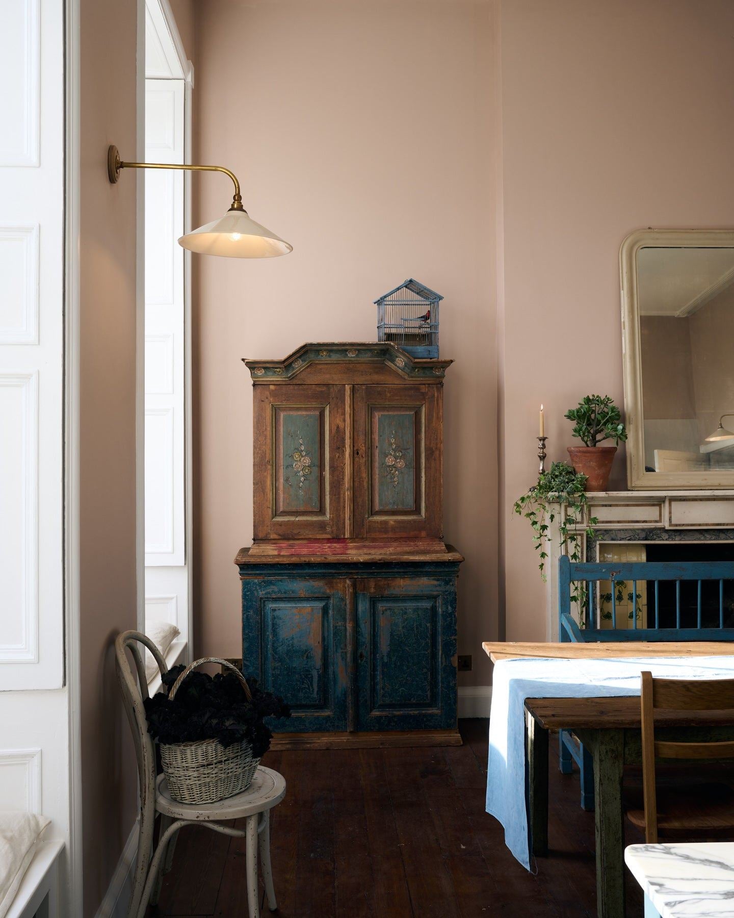 A grand room with high ceilings, soft pink walls, dark worn floorboards and antique furniture. A large Victorian-inspired wall light with a brass arm and a cream glazed ceramic shade is fixed to the wall by a tall window.