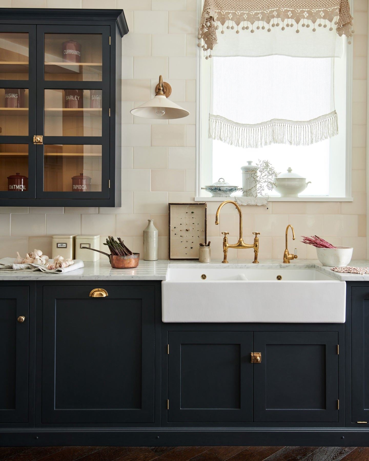 A straight-on view of a dark blue painted Shaker-style sink cupboard with a wide Belfast sink, an aged brass mixer tap, a wall of cream subway tiles and a large Victorian-inspired wall light with a brass arm and a cream glazed ceramic shade.
